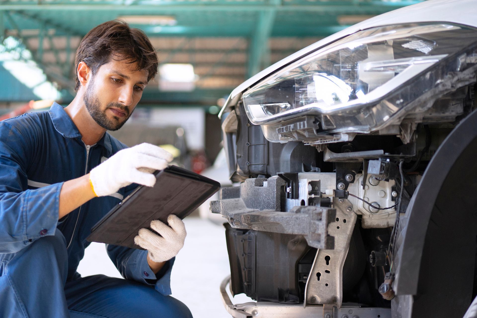 Mechanic in blue jumpsuit examines a car's damaged front end, using a tablet and wearing gloves, in a repair shop. Mechanic in blue jumpsuit examines a car's damaged front end, using a tablet and wearing gloves, in a repair shop.