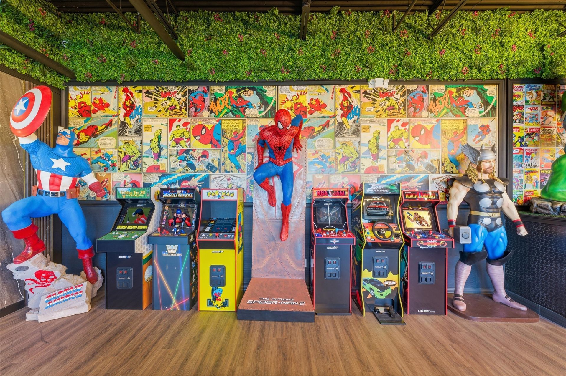Arcade with superhero statues: Captain America, Spider-Man, and Thor. Comic book wall art and multiple game consoles.
