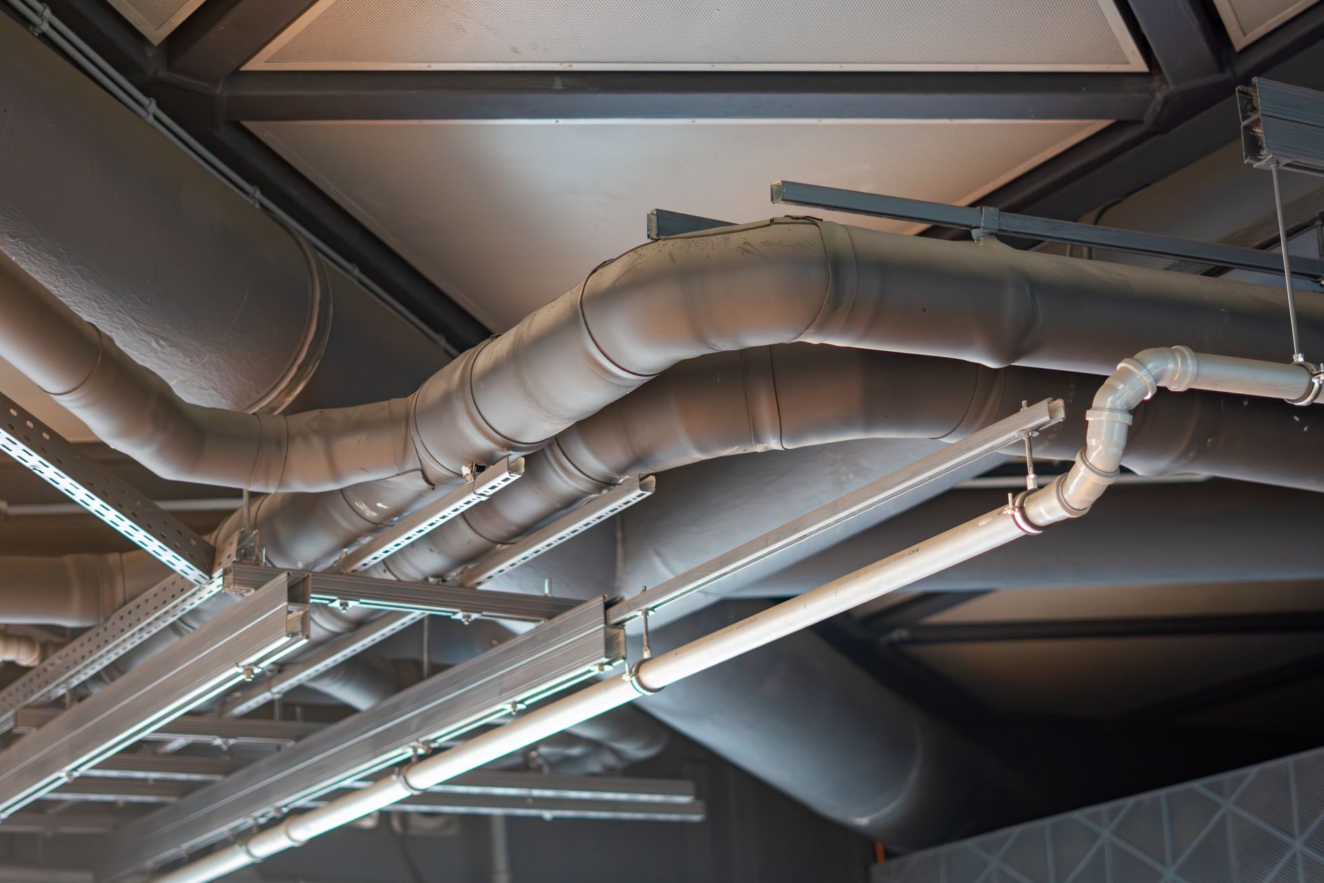 Ceiling view of exposed silver HVAC ducts and fluorescent lights in an industrial interior