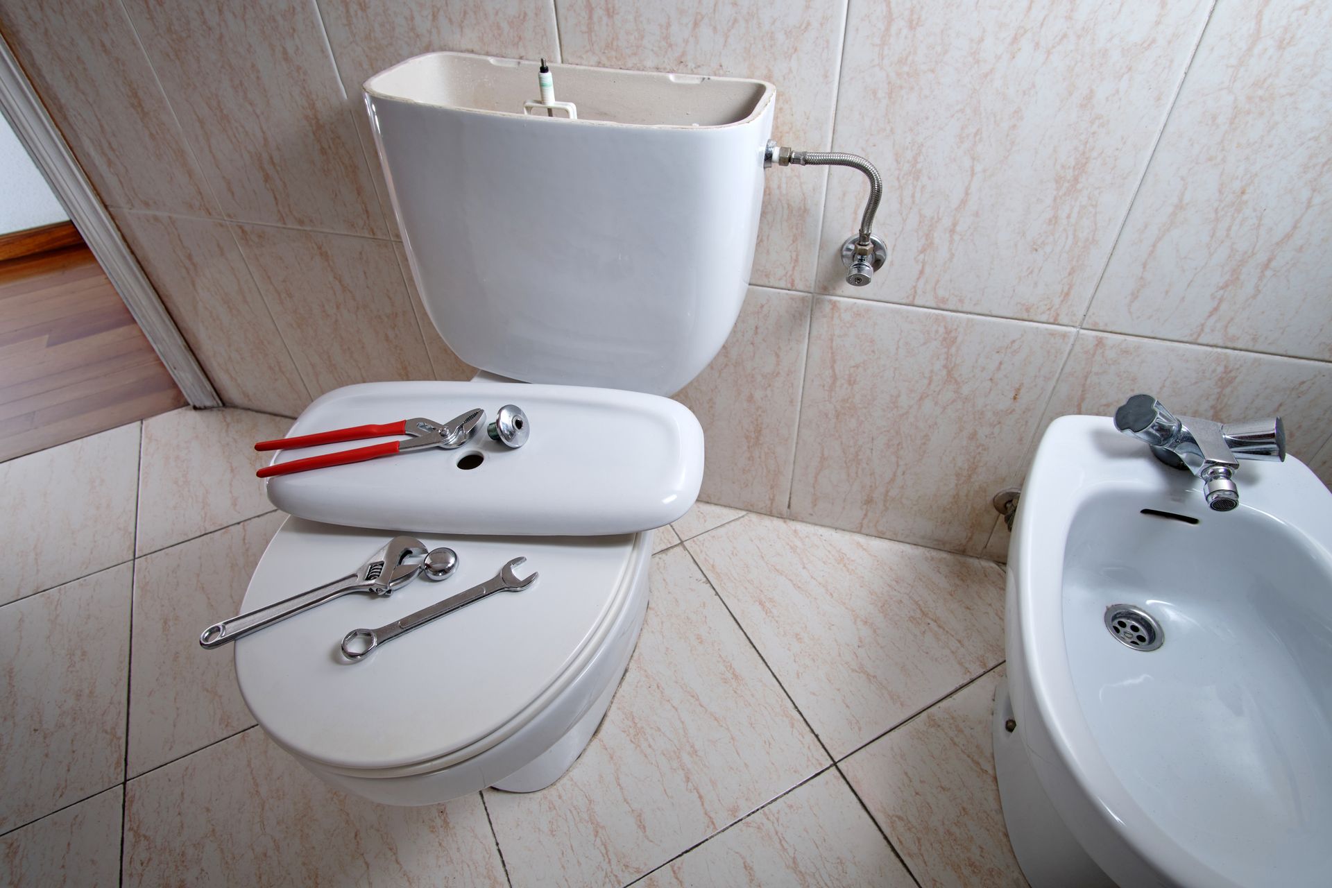 A toilet with its tank lid removed and repair tools placed on the seat in a tiled bathroom next to a bidet.