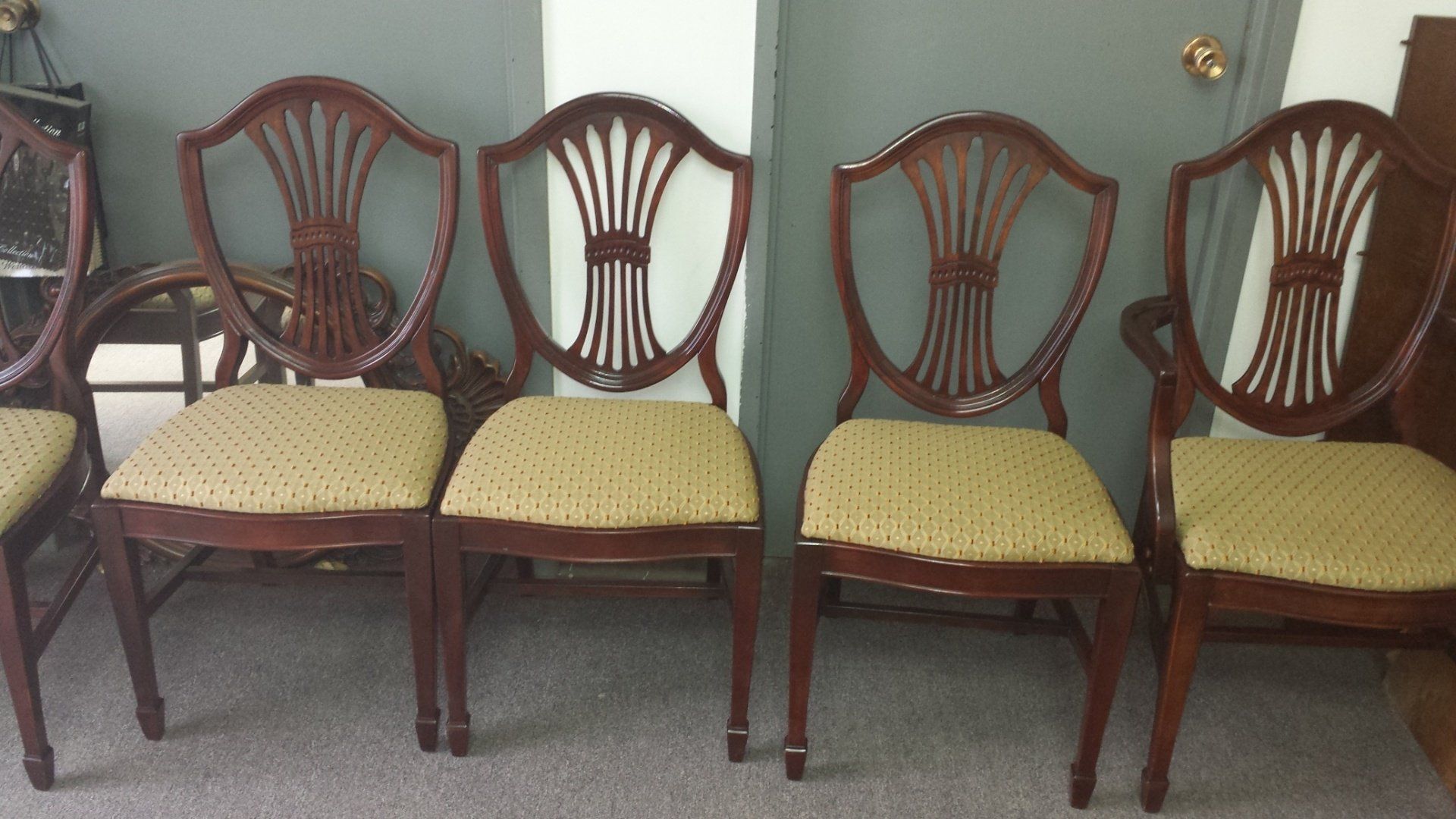 Four wooden chairs with yellow seats lined up against a teal wall on a carpeted floor.