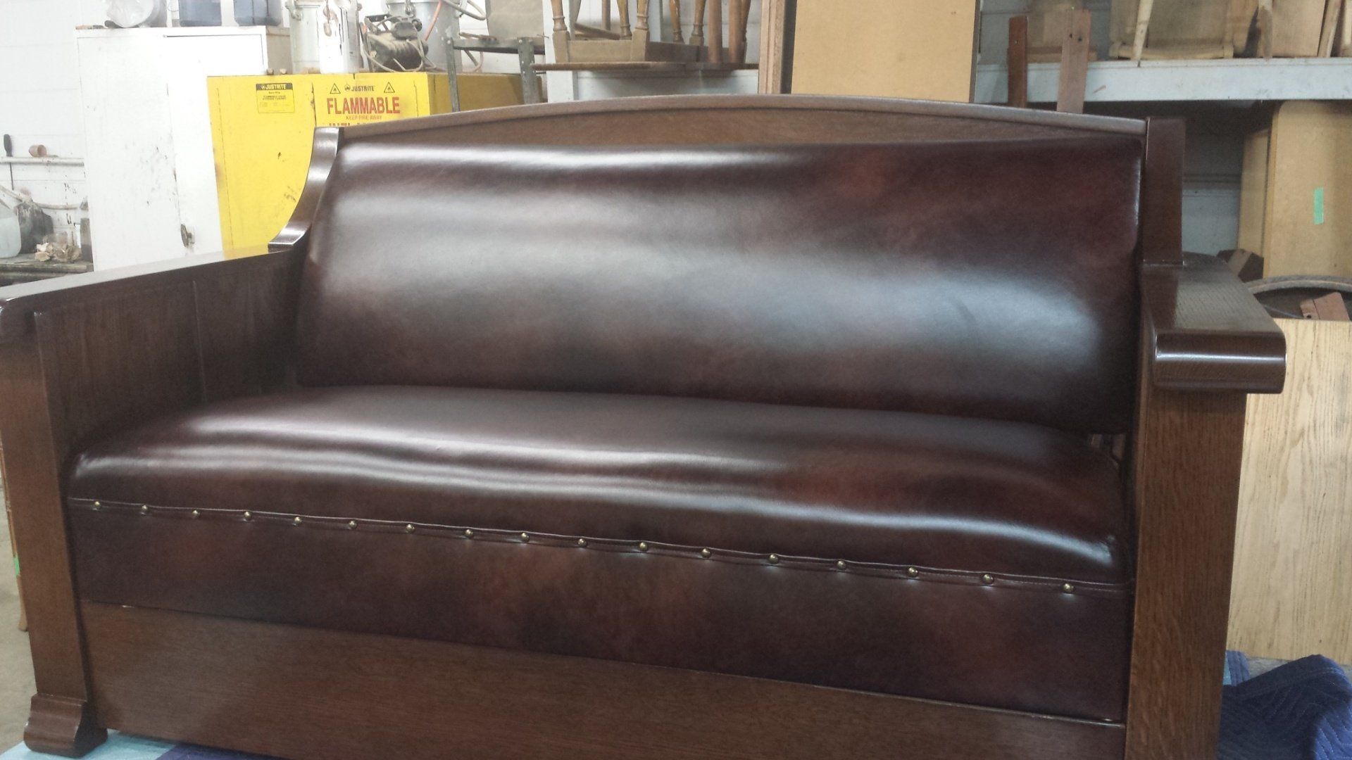 Dark brown leather loveseat in a workshop, with wooden arms and backrest.