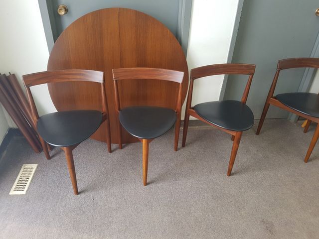 Four wooden chairs with black seats in a room, with a round wooden tabletop leaning behind them