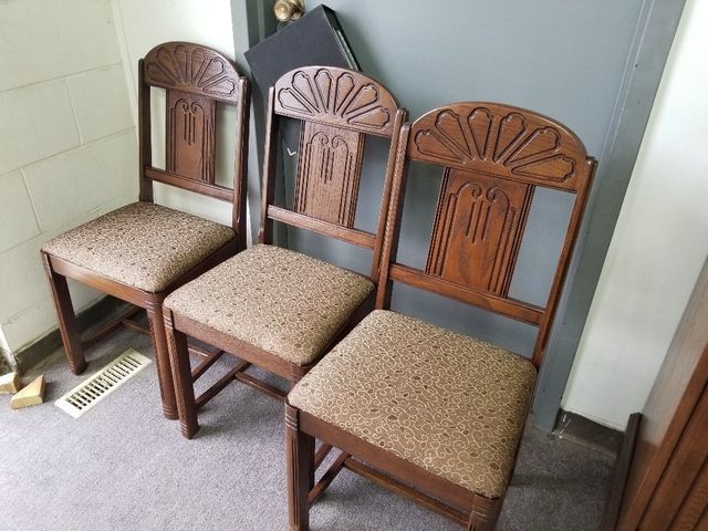 Three wooden chairs with beige cushions lined up against a wall in a corner.