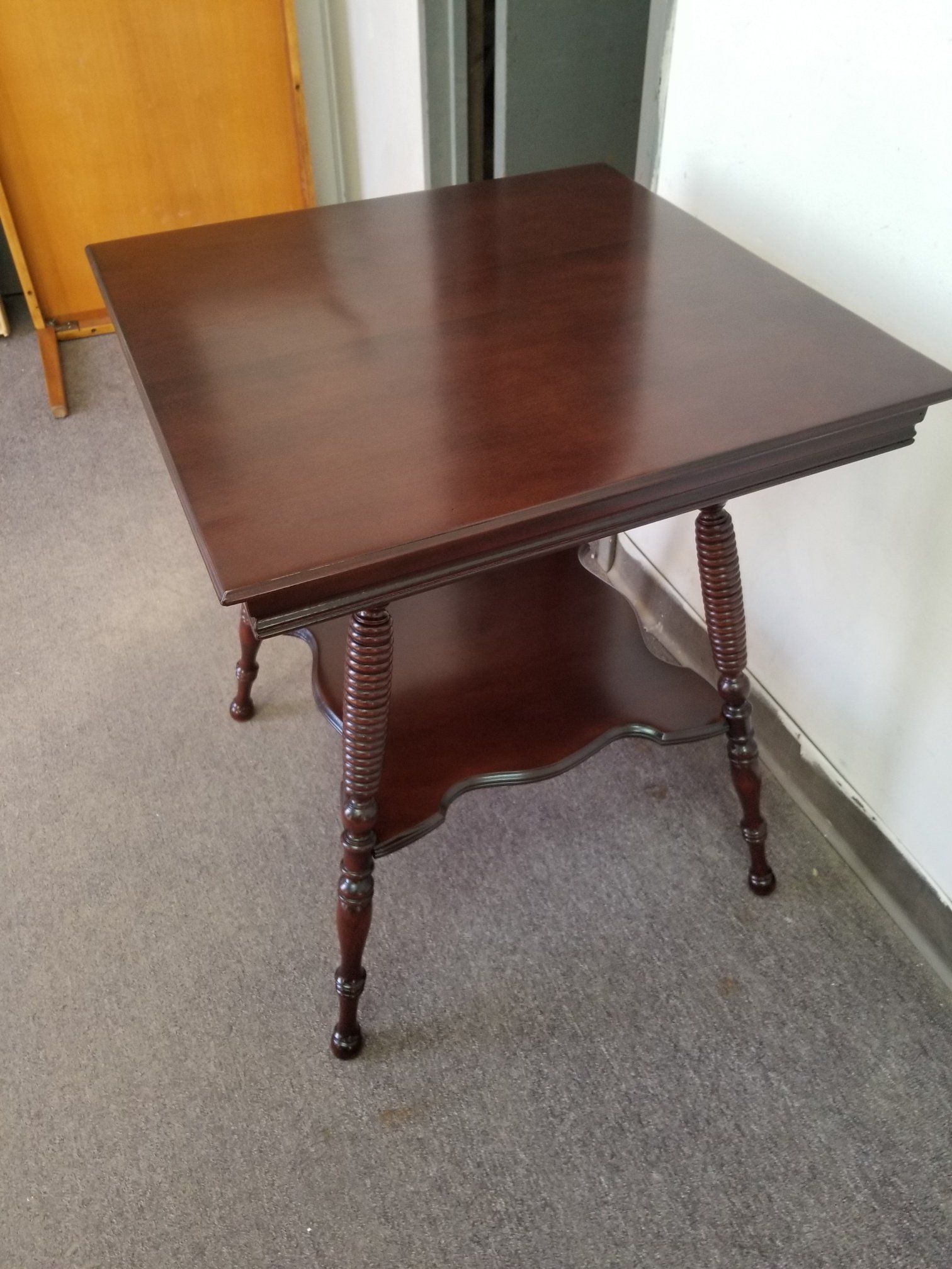Dark wooden side table with turned legs and lower shelf on carpet beside a wall