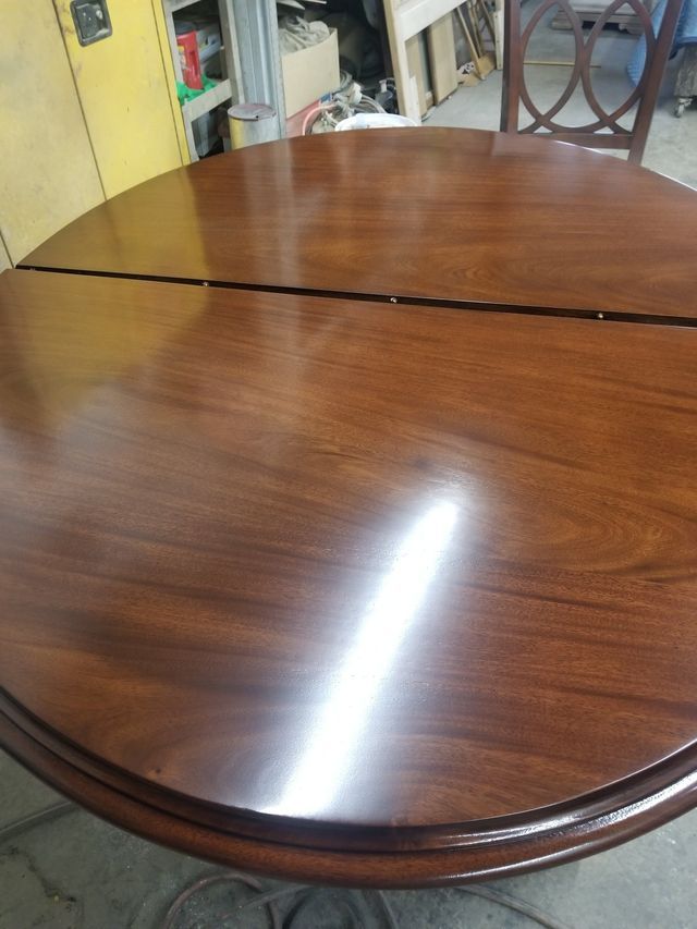 Round wooden dining table with a visible seam, in a room with chairs and shelves in the background