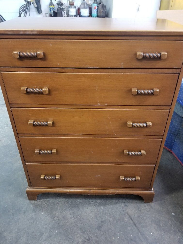 Wooden five-drawer dresser with brass handles on a carpeted floor, bottles visible on top.