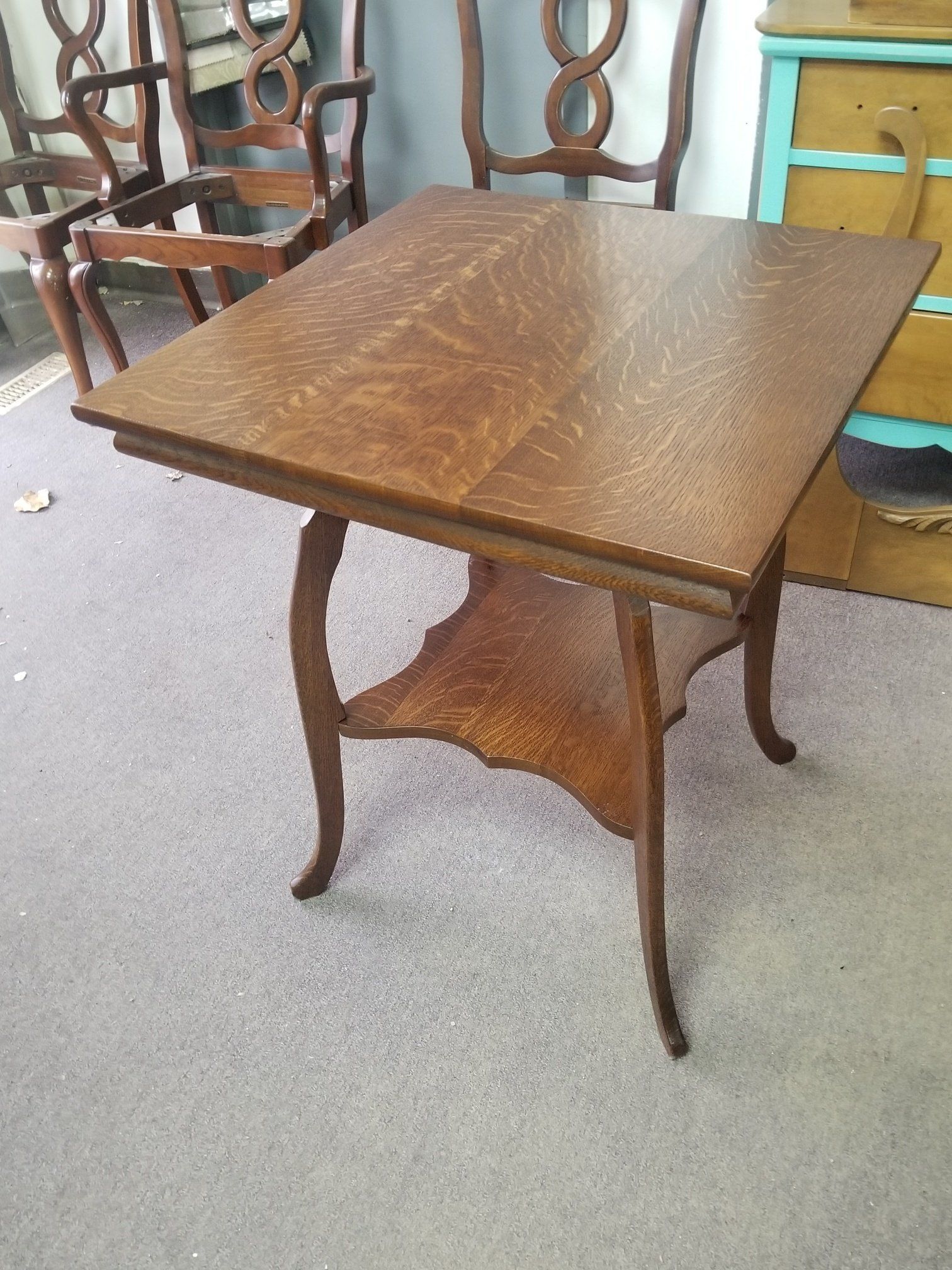 Small wooden square table with curved legs in a carpeted room, with chairs and a teal cabinet behind it