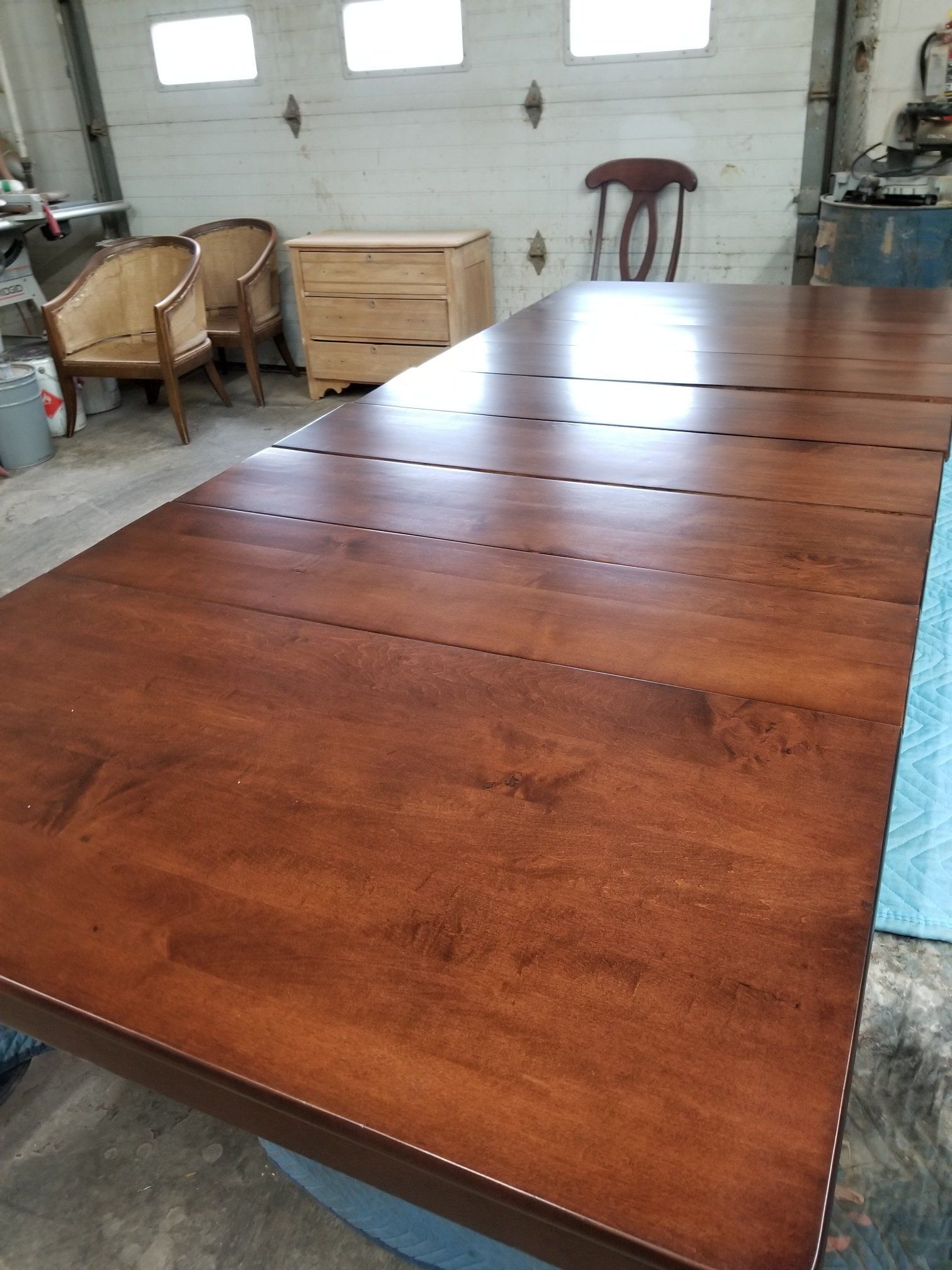 Large polished wooden table in a workshop, with chairs and storage crates in the background.
