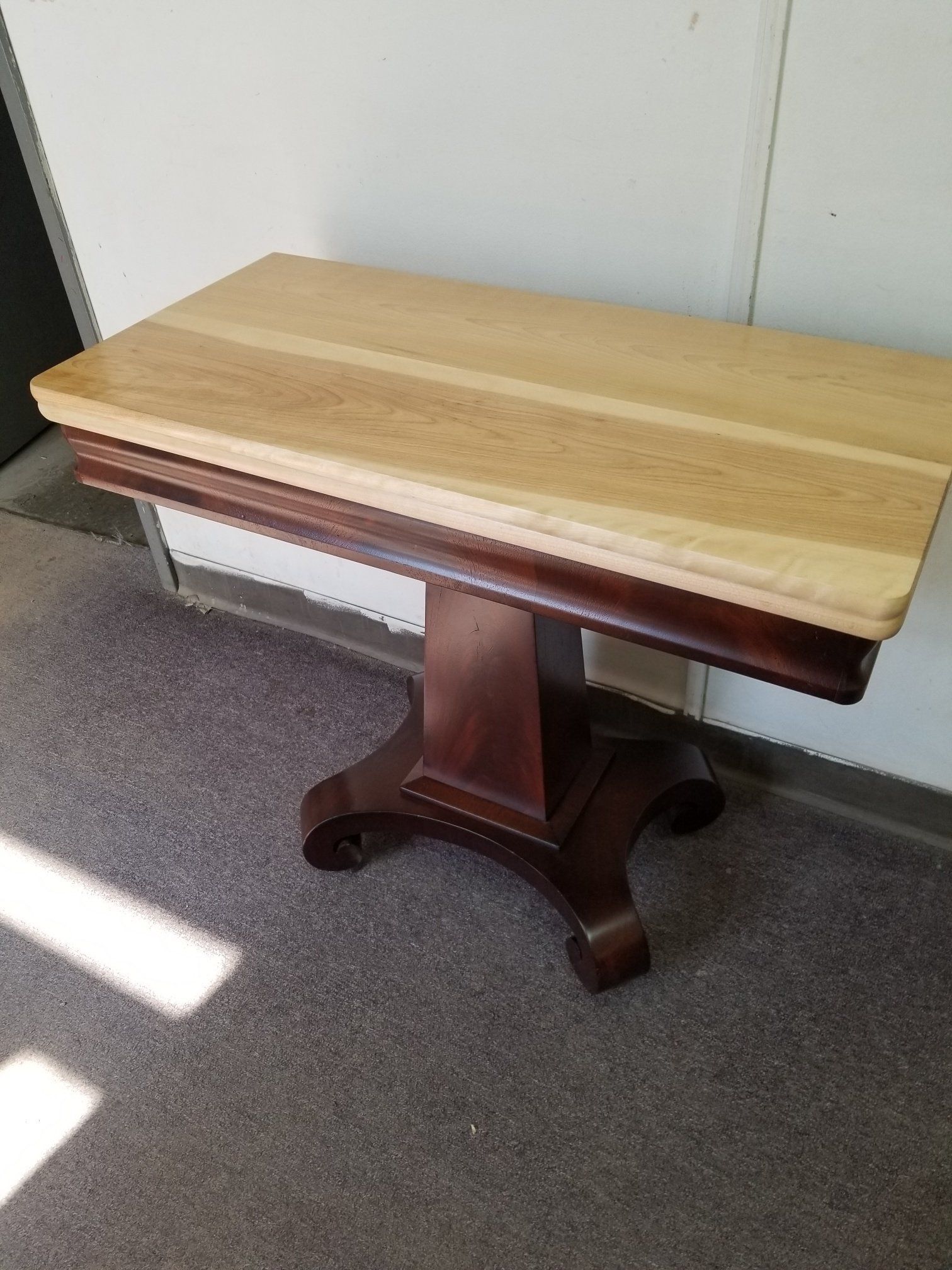 Wooden table with a light top and dark pedestal base in a carpeted room.