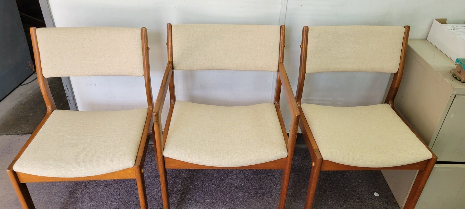 Three wooden chairs with beige seats and backs lined up against a wall.
