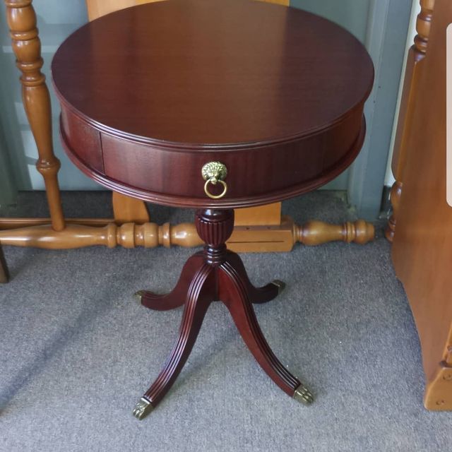 Small round dark wood side table with brass drawer pull and tripod base on carpet.