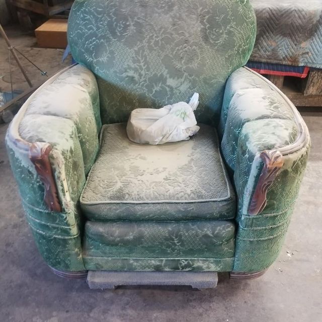 Dusty green upholstered armchair with worn arms and a white cloth on the seat in a workshop or storage area