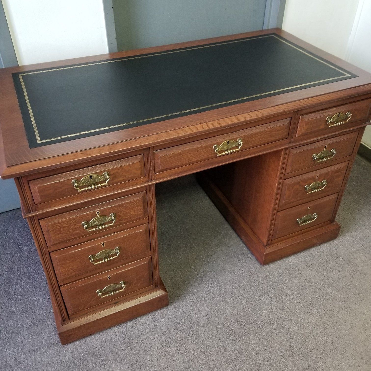 Wooden executive desk with black leather top and brass drawer pulls, shown in a carpeted room.