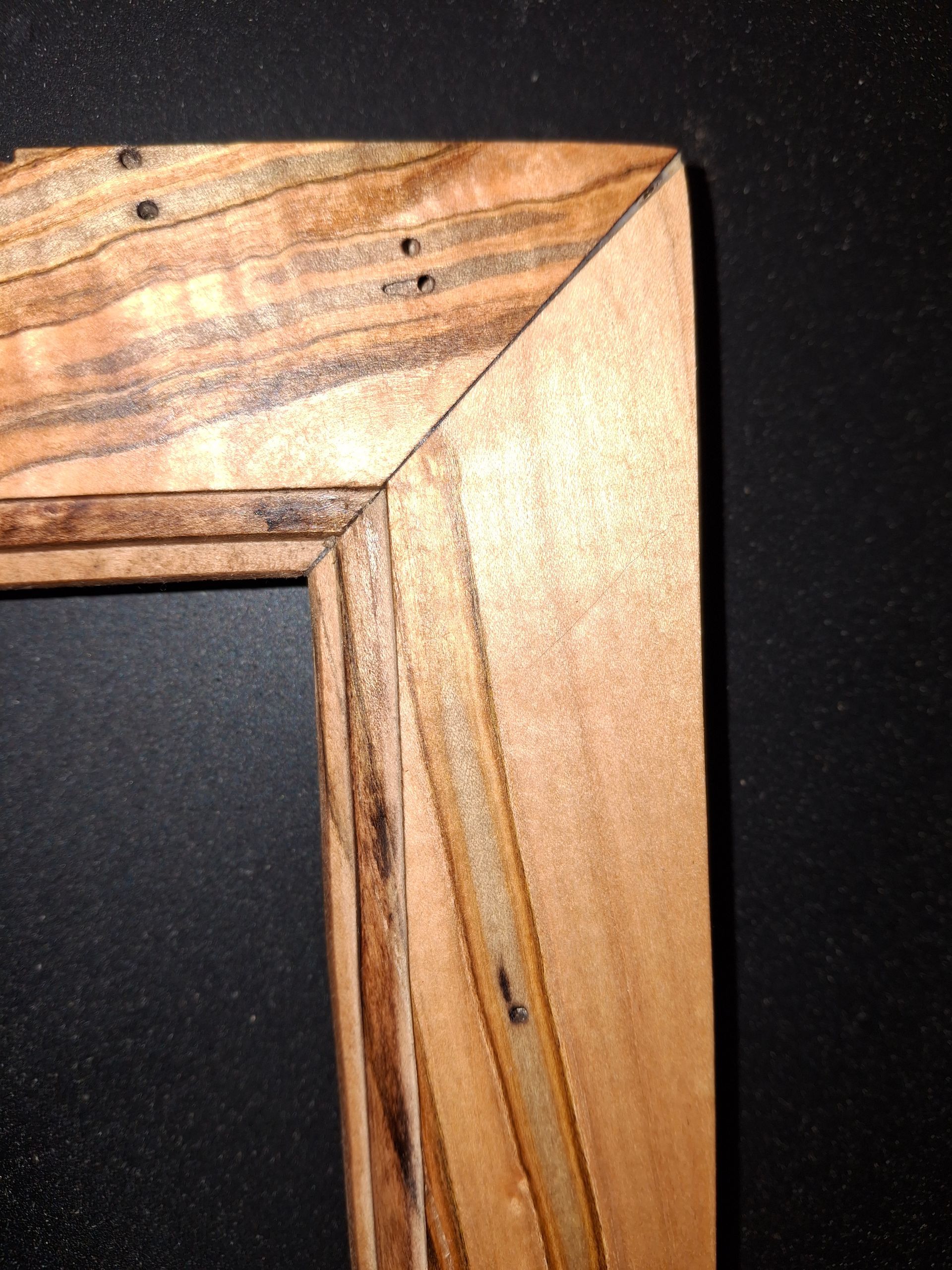 A corner detail of a wooden picture frame with natural grain patterns and visible pinholes at the mitered joint.