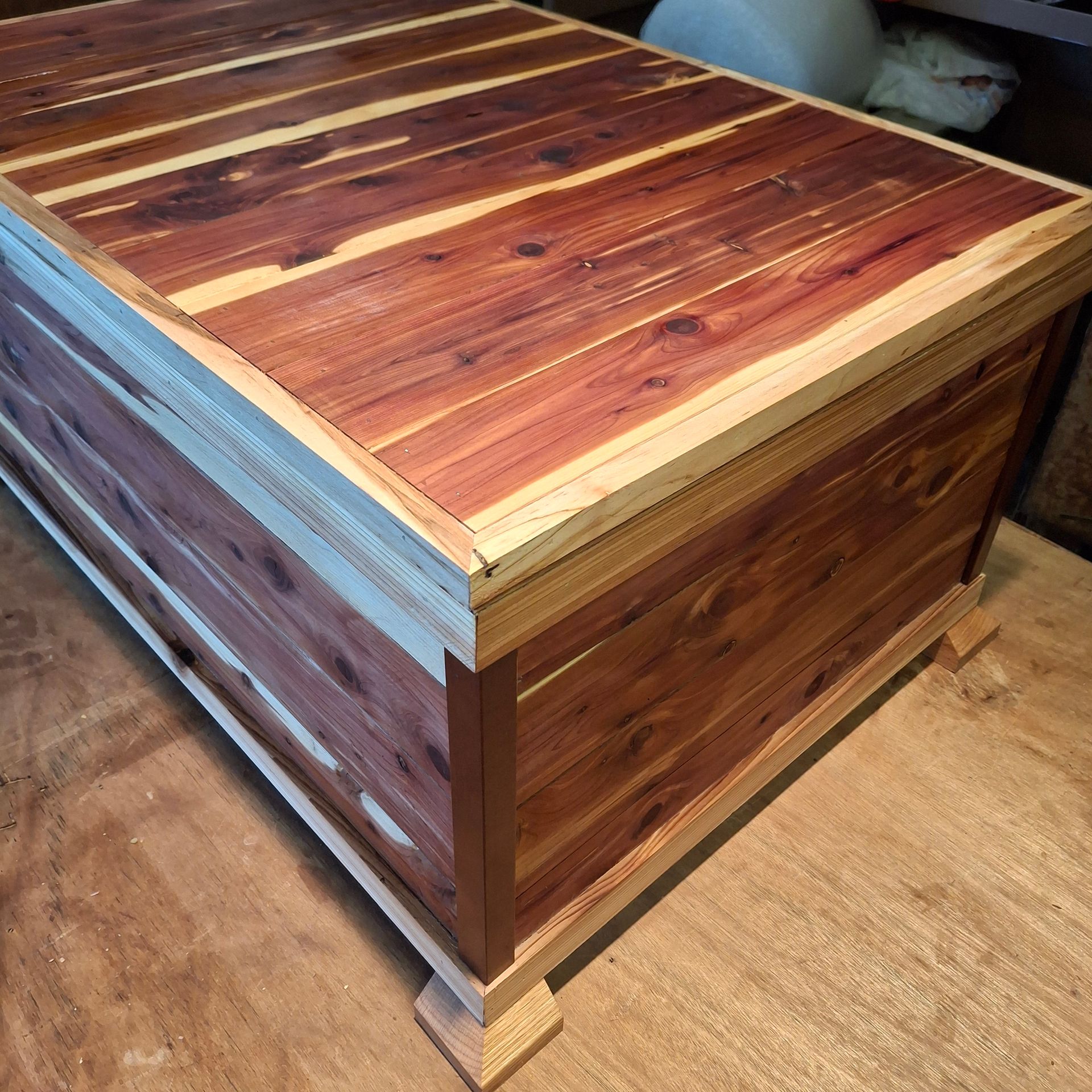 A rustic cedar wood chest with a hinged lid, featuring alternating red and blonde wood grain panels on a corner footed base.
