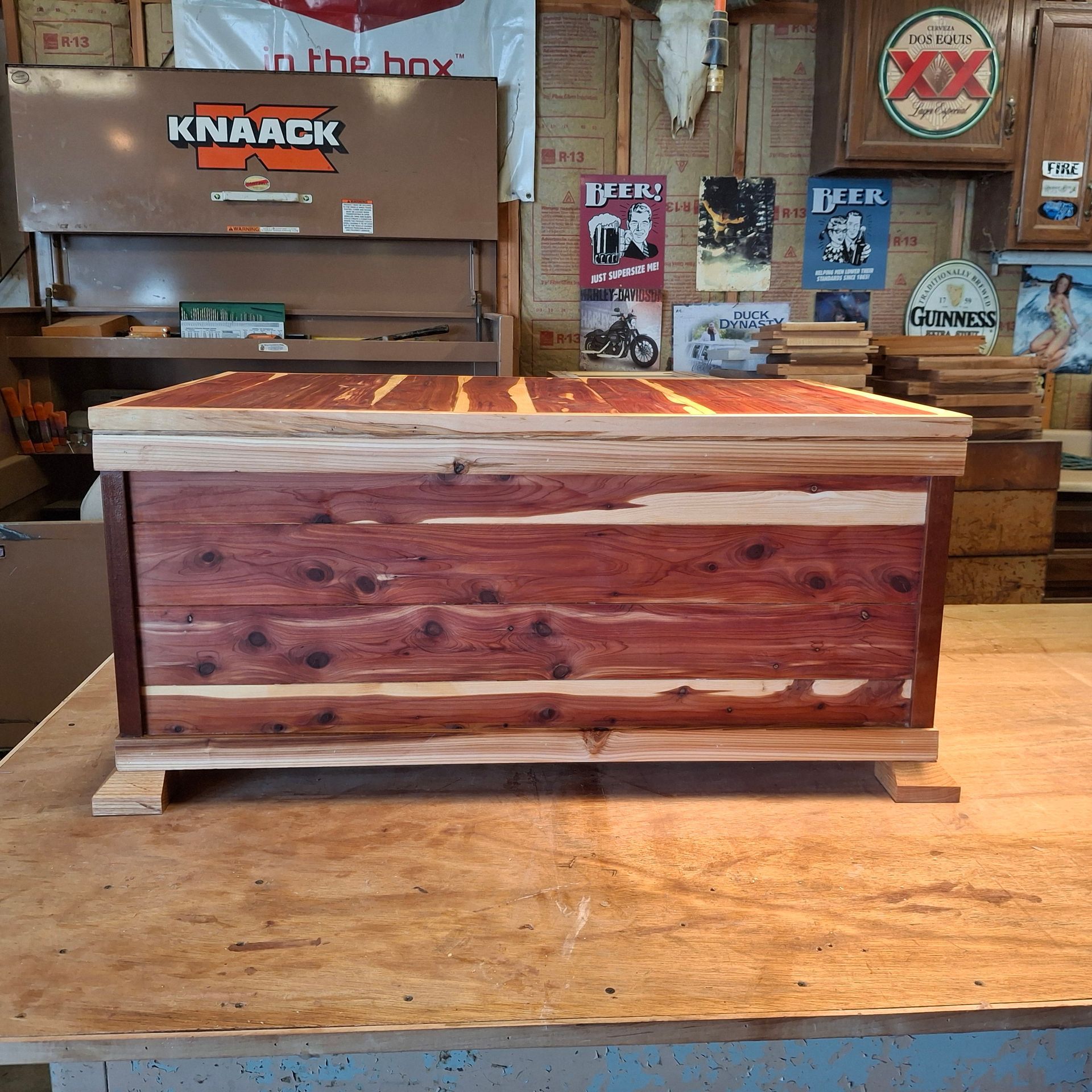 A handmade wooden cedar chest sits on a workbench in a workshop filled with tools, signs, and woodworking supplies.