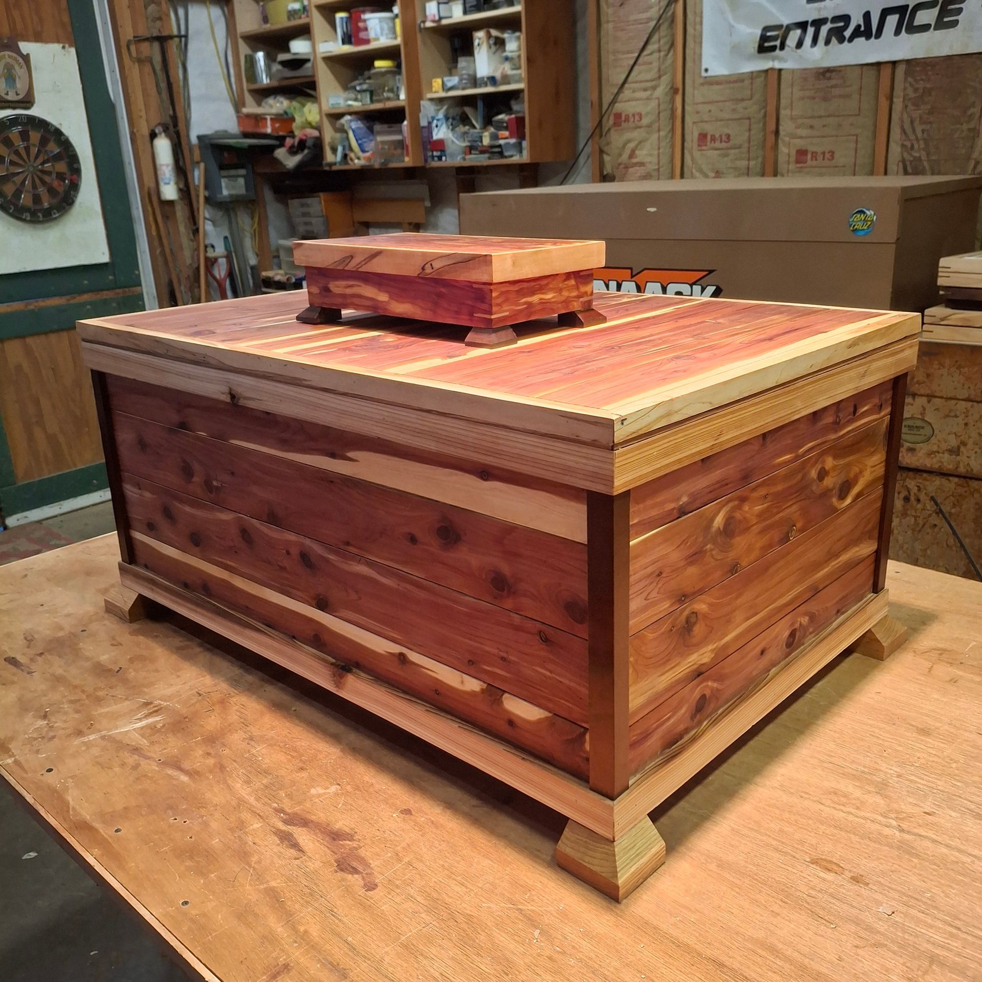 A handmade red cedar chest with lighter trim and a small matching box on top, sitting on a wooden workbench in a shop.