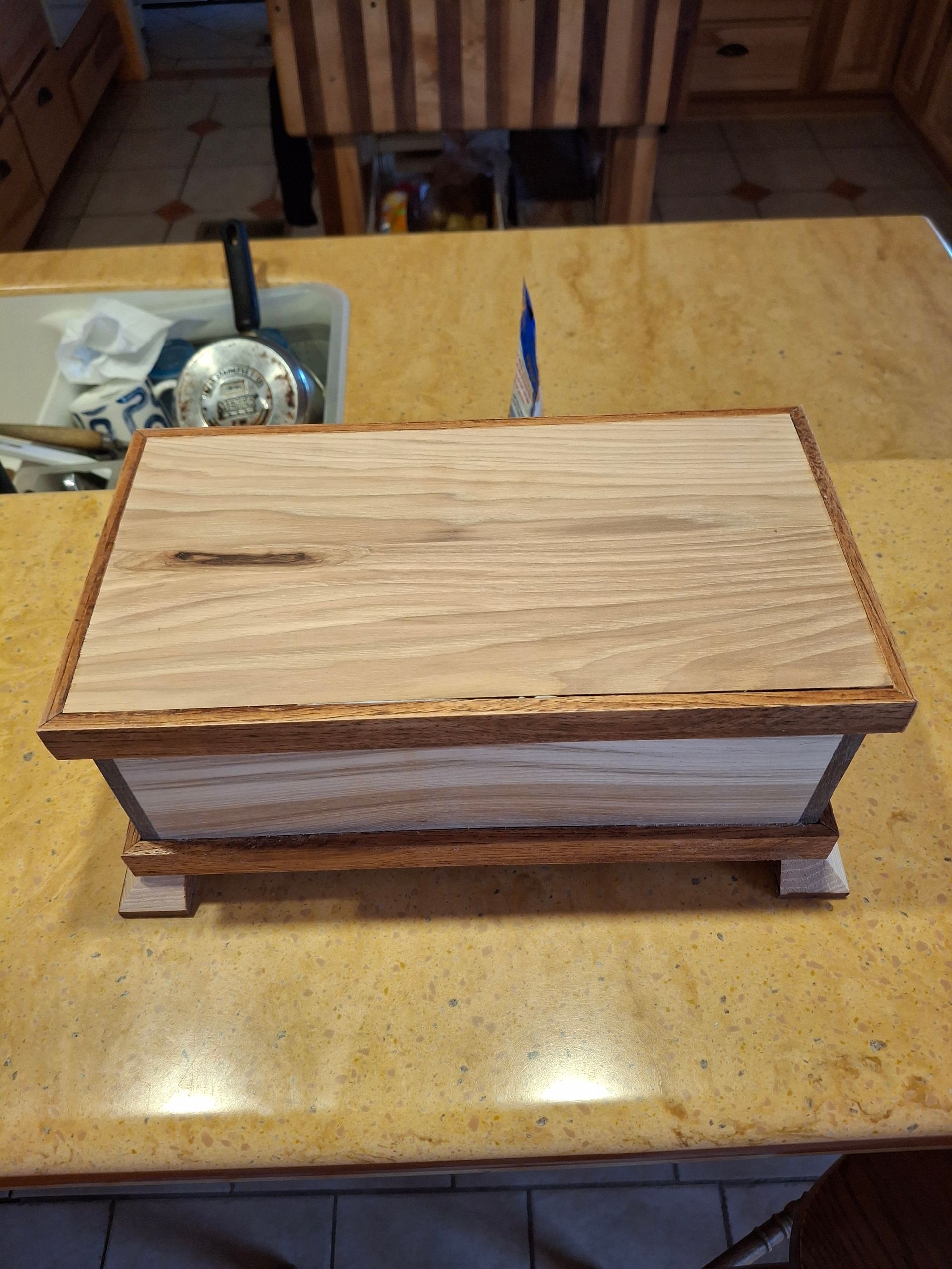 A wooden box with a textured, dark-edged trim and a lighter wood lid featuring a natural knot, sitting on a countertop.