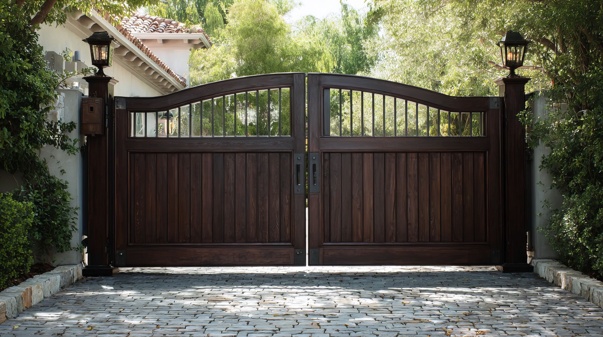 Double wooden driveway gates with a dark finish, arched tops, and metal grilles, flanked by stone pillars and lanterns.
