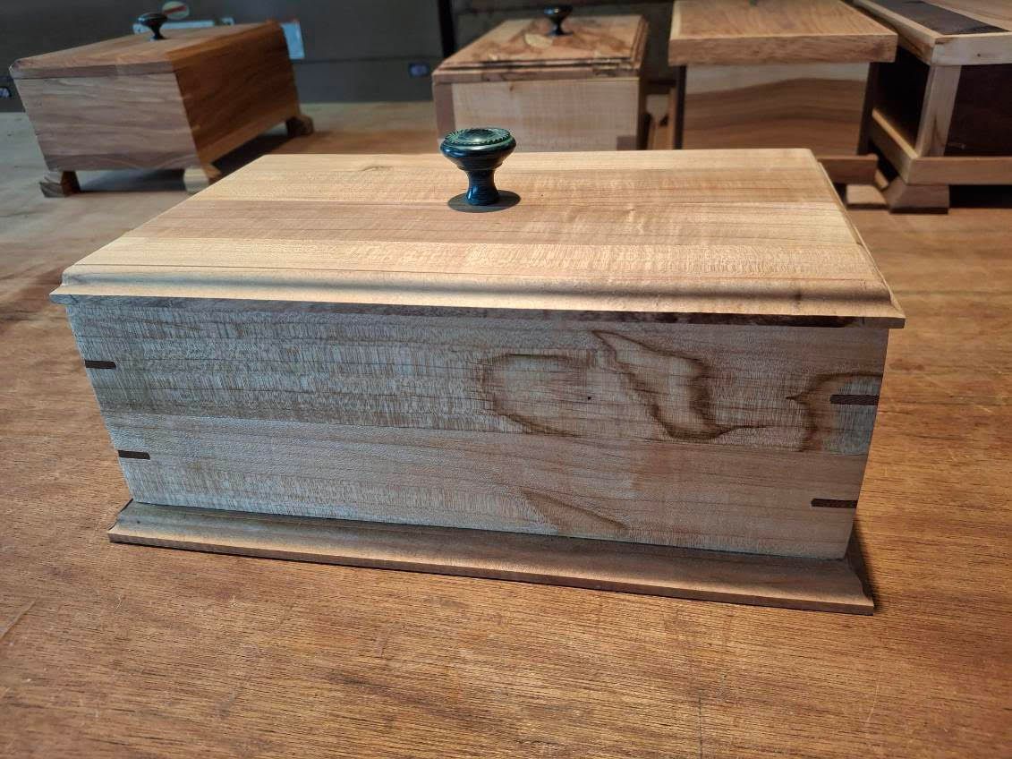 Wooden keepsake box with a lid and dark knob, displayed on a table in a workshop or showroom.