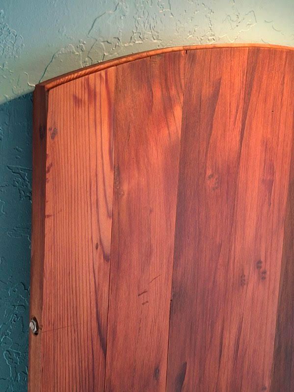 Close-up of a reddish wooden door or panel with a curved top against a pale wall.