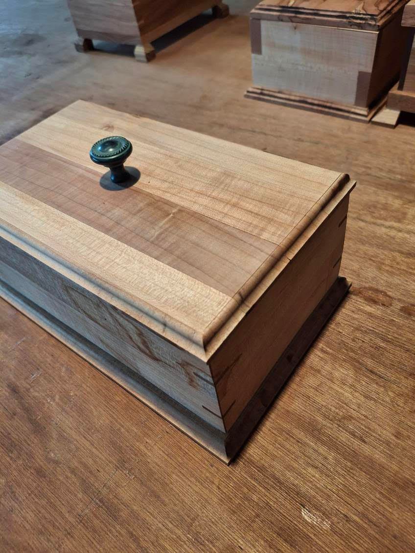 Small wooden box with a dark knob on top, sitting on a wooden floor in a workshop or storage area.