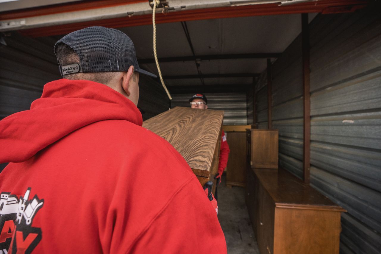 Two men in a storage unit carrying a wooden board. One in a red hoodie is in the foreground.