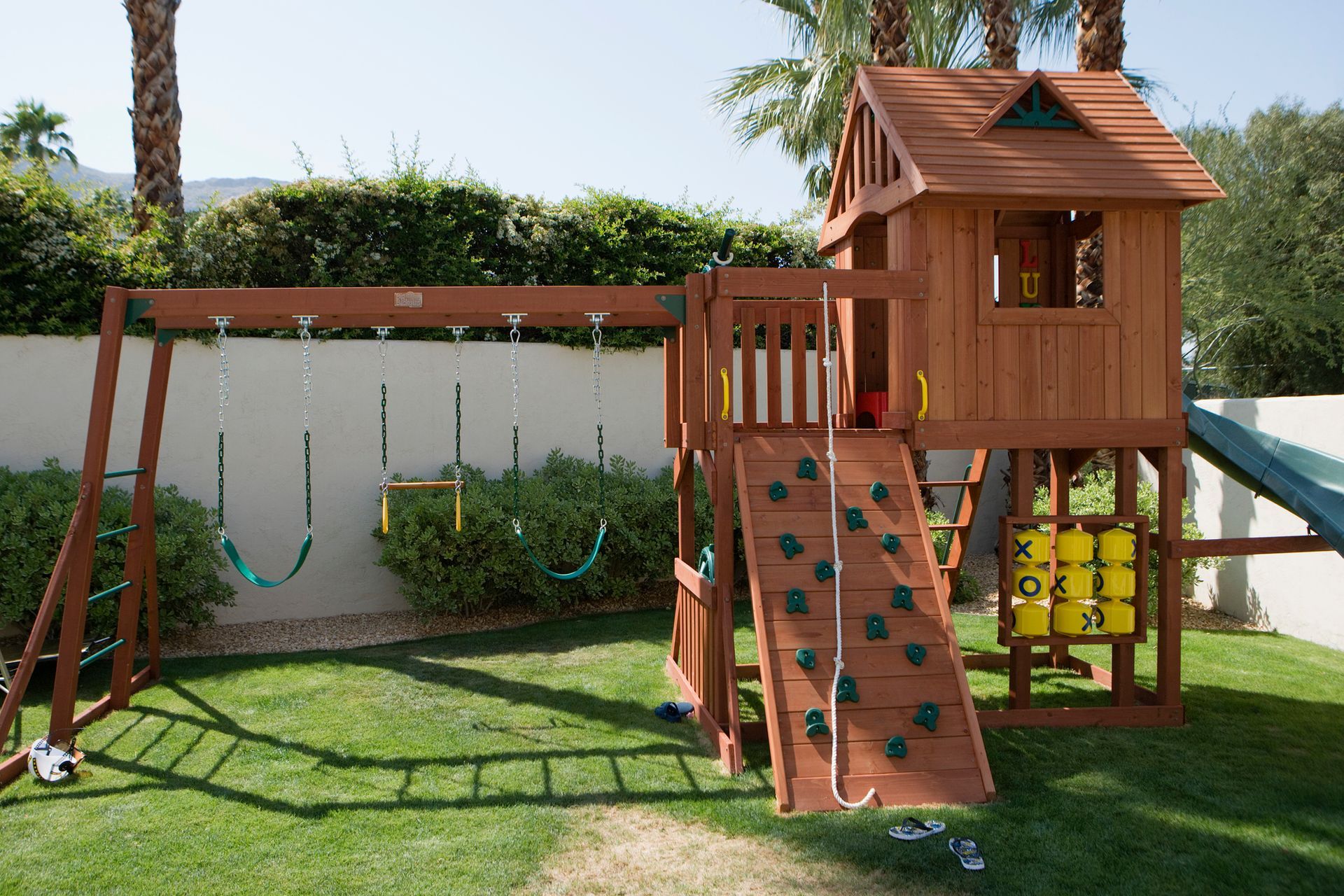 Wooden playset with swings, climbing wall, slide, and playhouse on green grass.