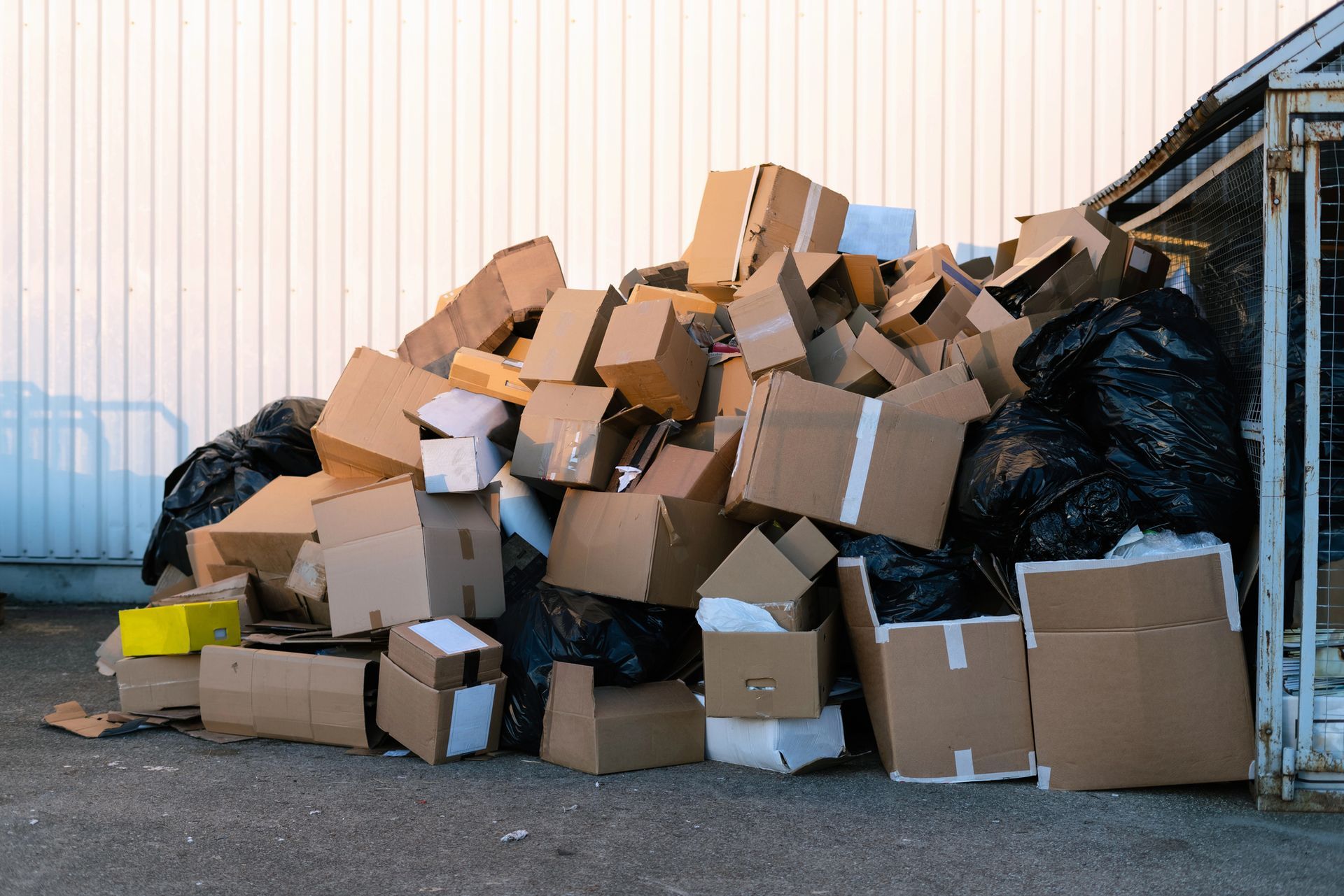 Pile of cardboard boxes and black trash bags next to a white building and a metal cage.