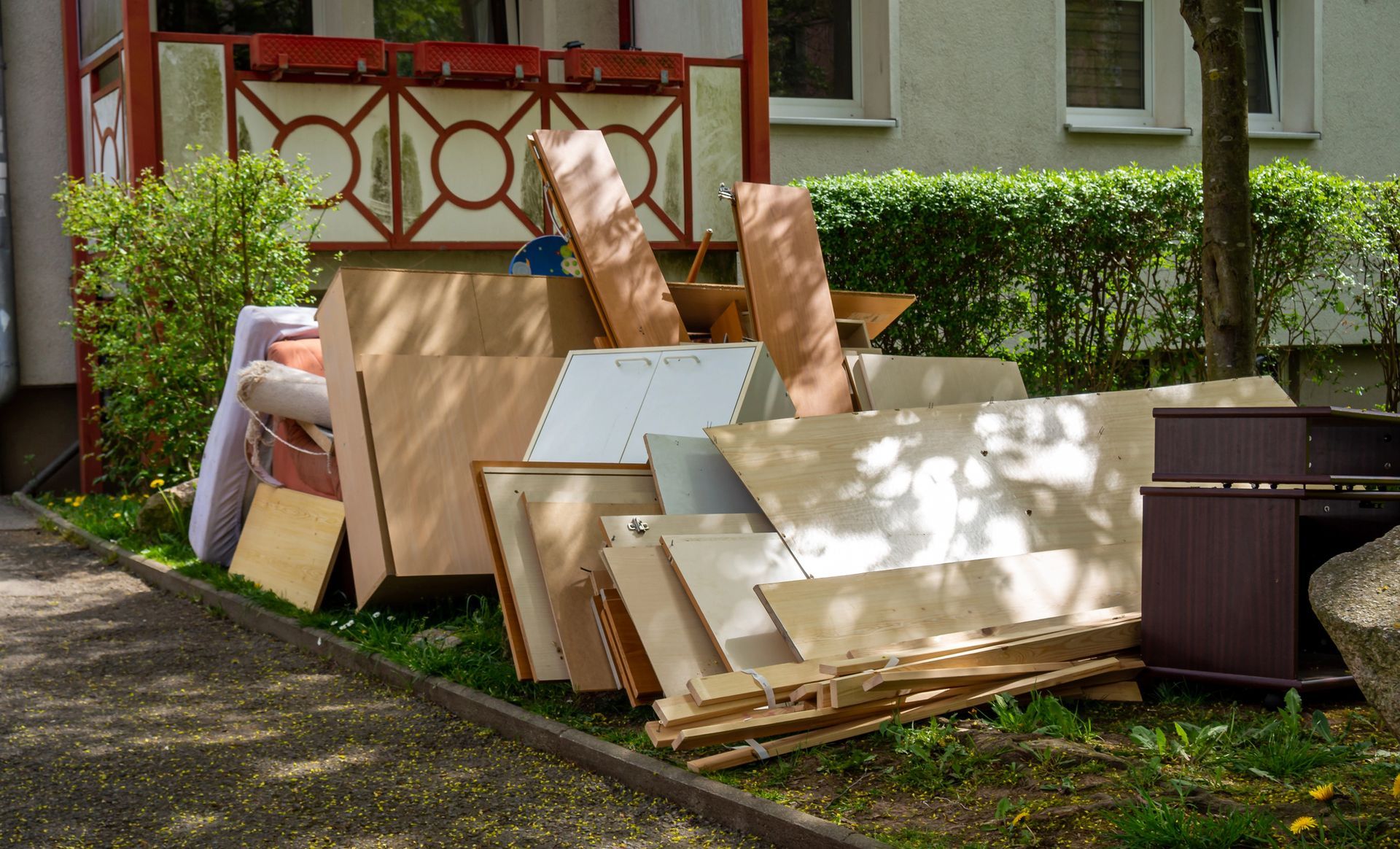 Pile of discarded wooden furniture and panels on a grassy area near a building.