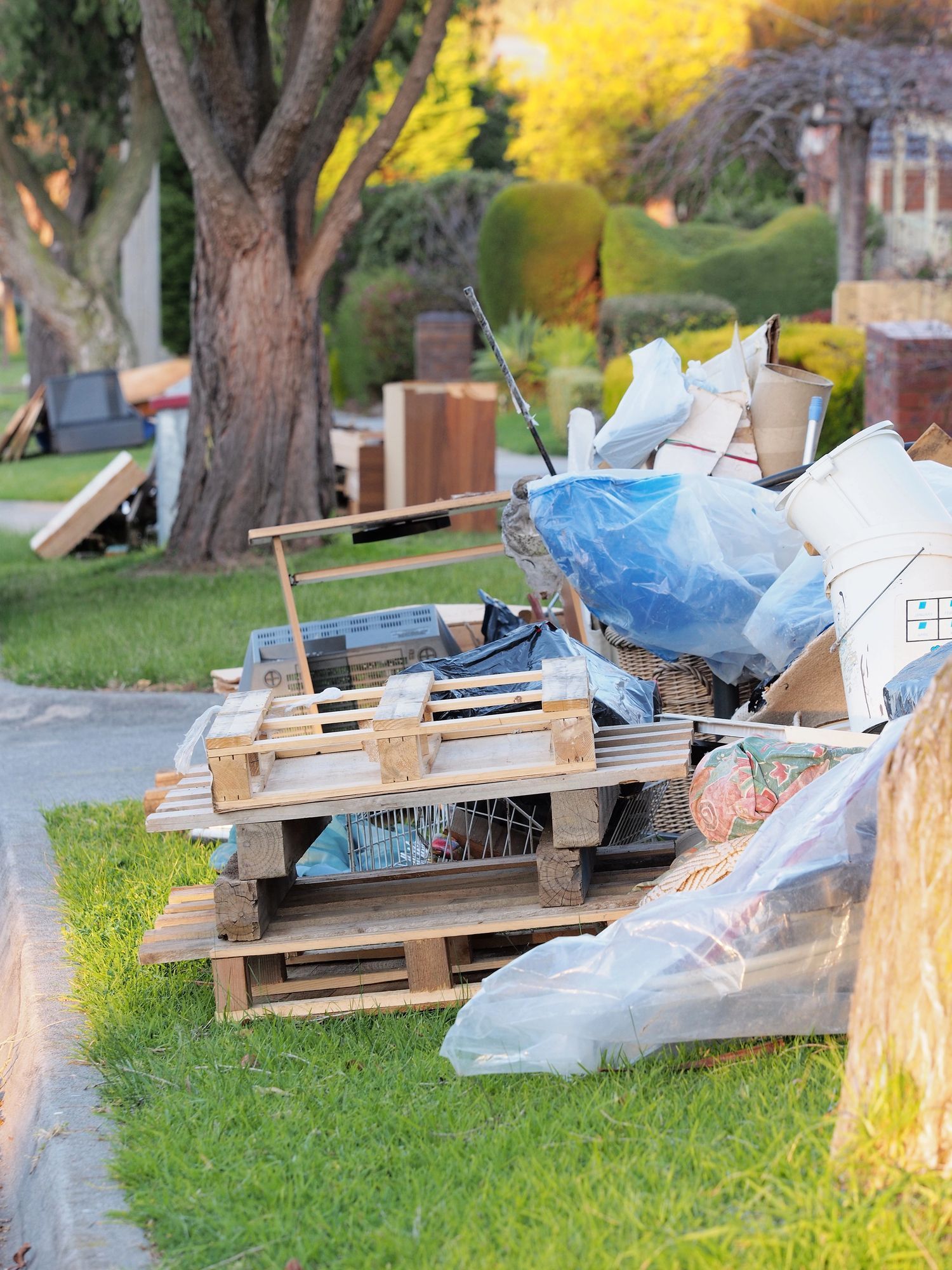 Pile of discarded items on a lawns in a neighborhood, including pallets, furniture, and plastic-wrapped waste.