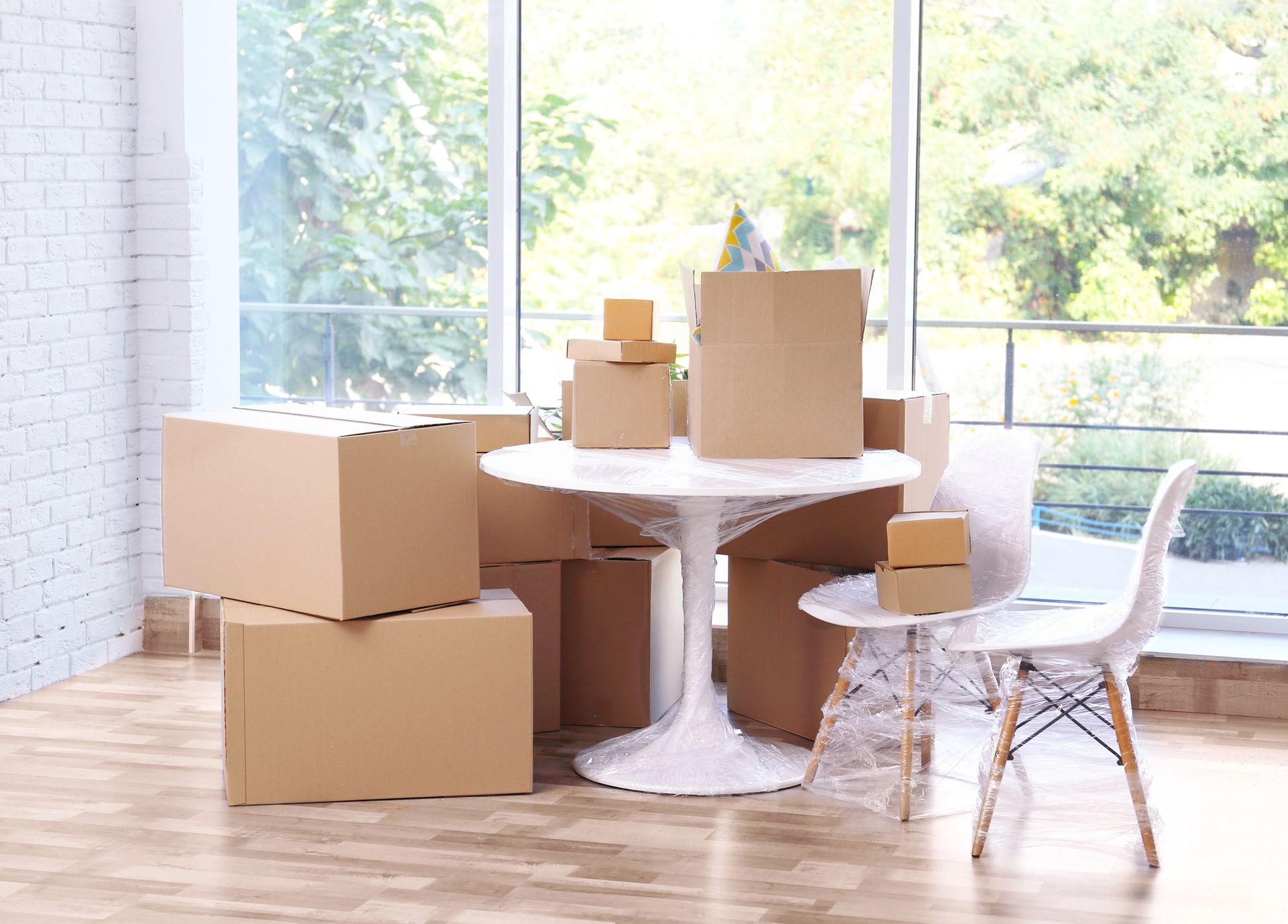 Cardboard boxes on and around a white table with a chair, near a large window with a tree view.