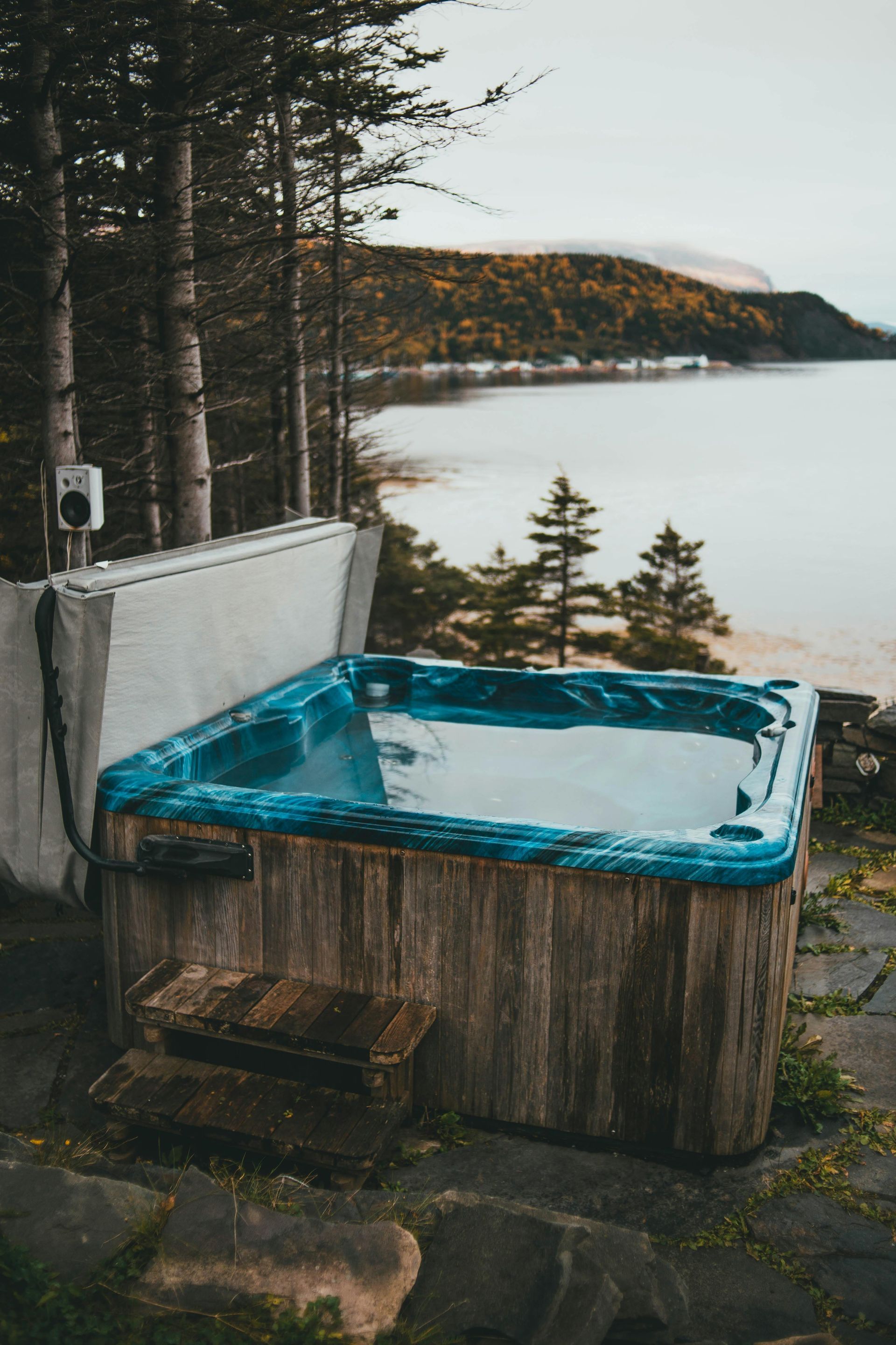 Hot tub overlooking a lake, set in a forest. Wooden tub with blue interior, overcast sky.