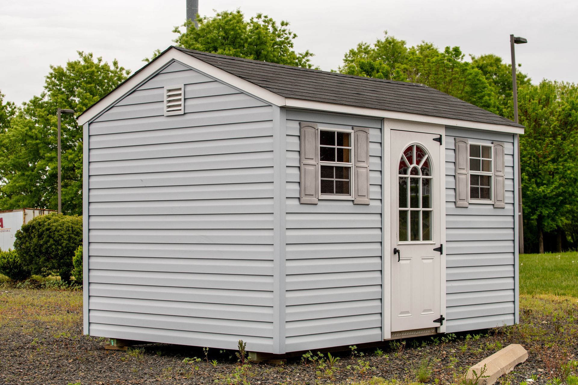 Light blue shed with white door and two small windows, set in gravel, with green trees in the background.