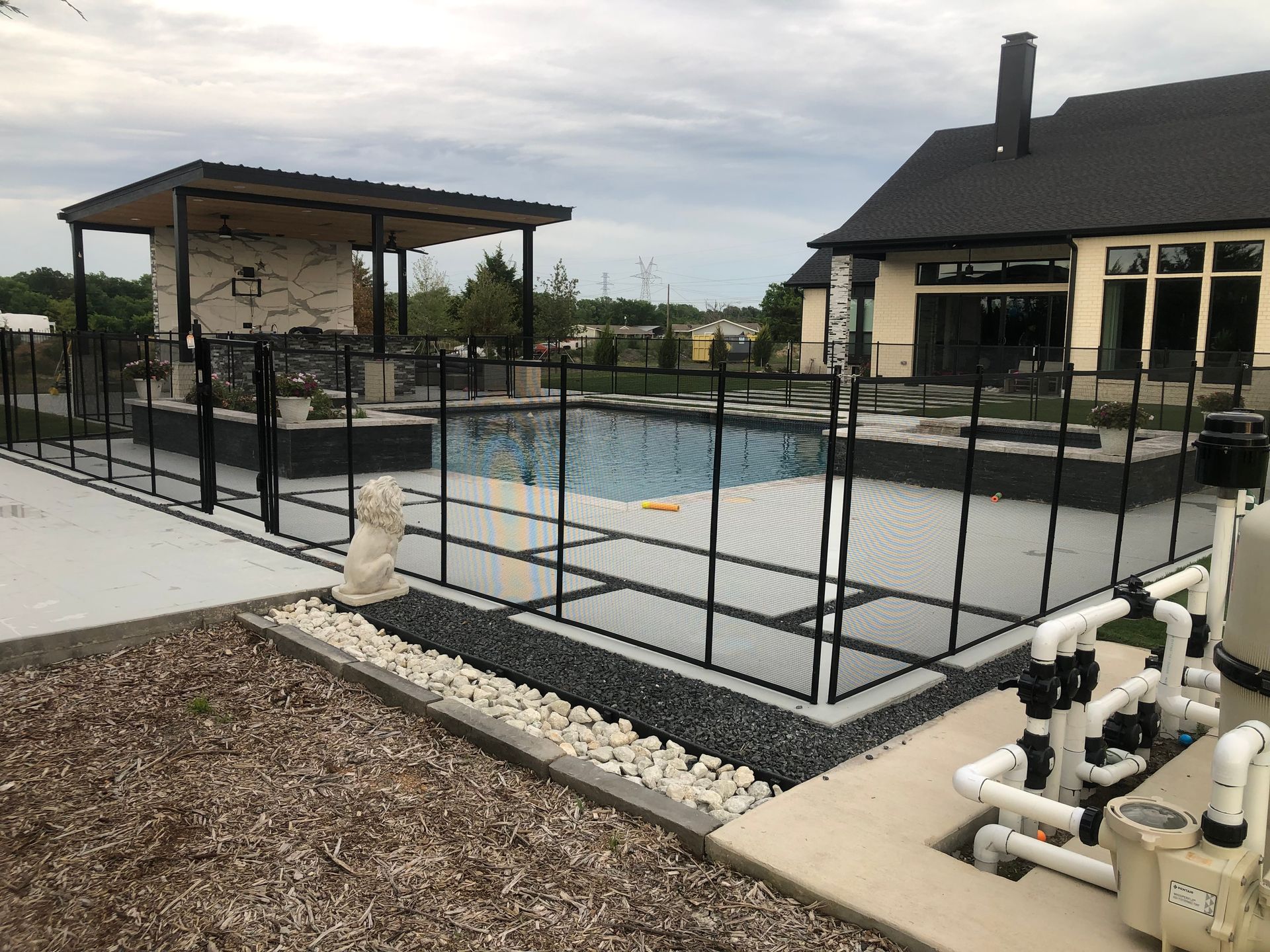 Outdoor pool area with black safety fence, covered patio, and adjacent house under cloudy sky