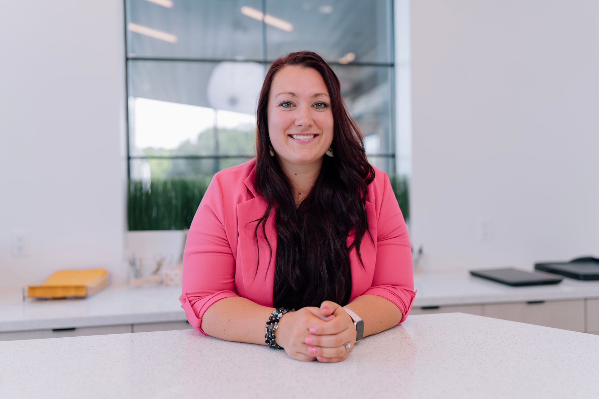 Woman in a pink blazer seated at a white counter in a bright office, smiling at the camera.