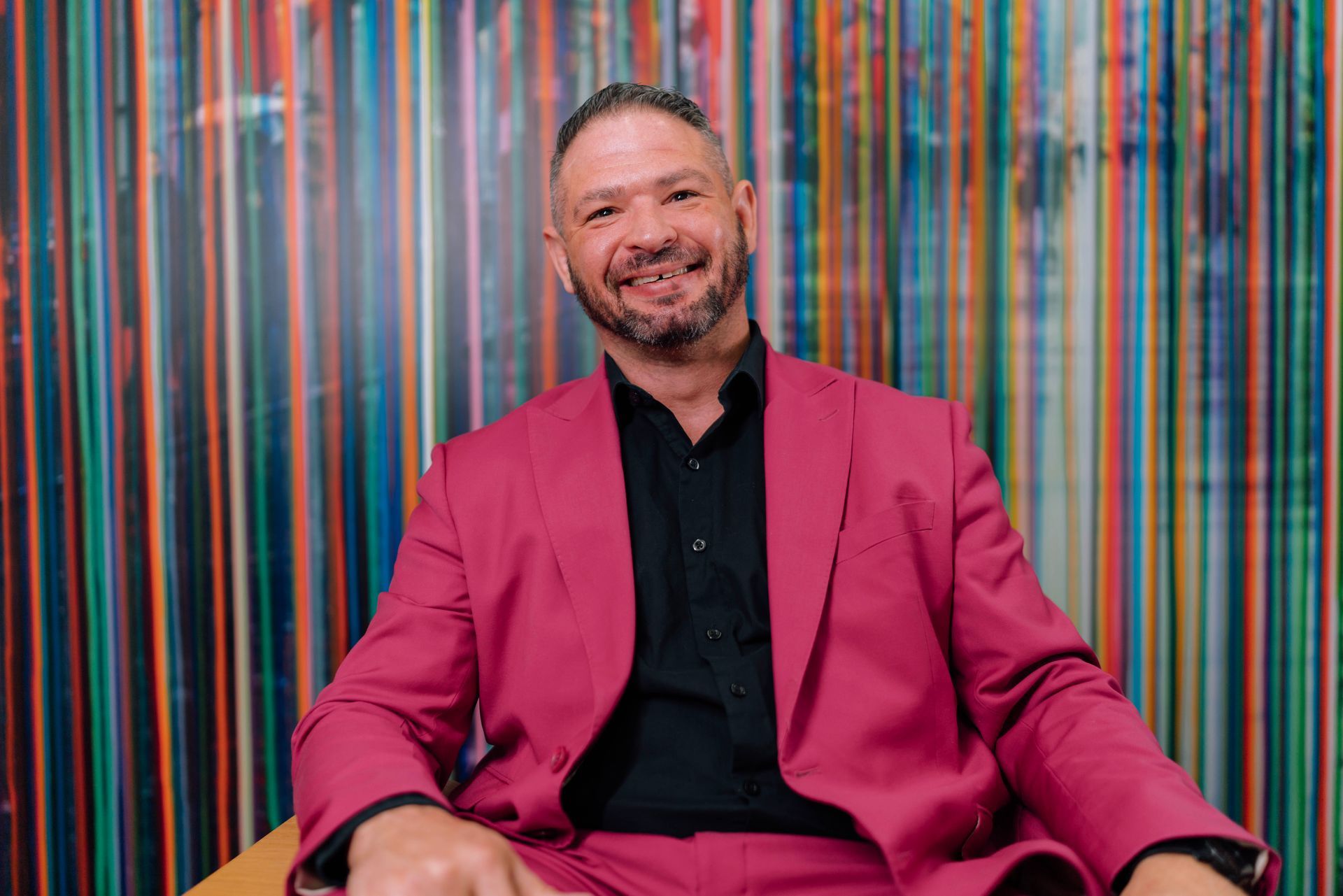 Smiling man in a magenta suit seated before a colorful striped backdrop
