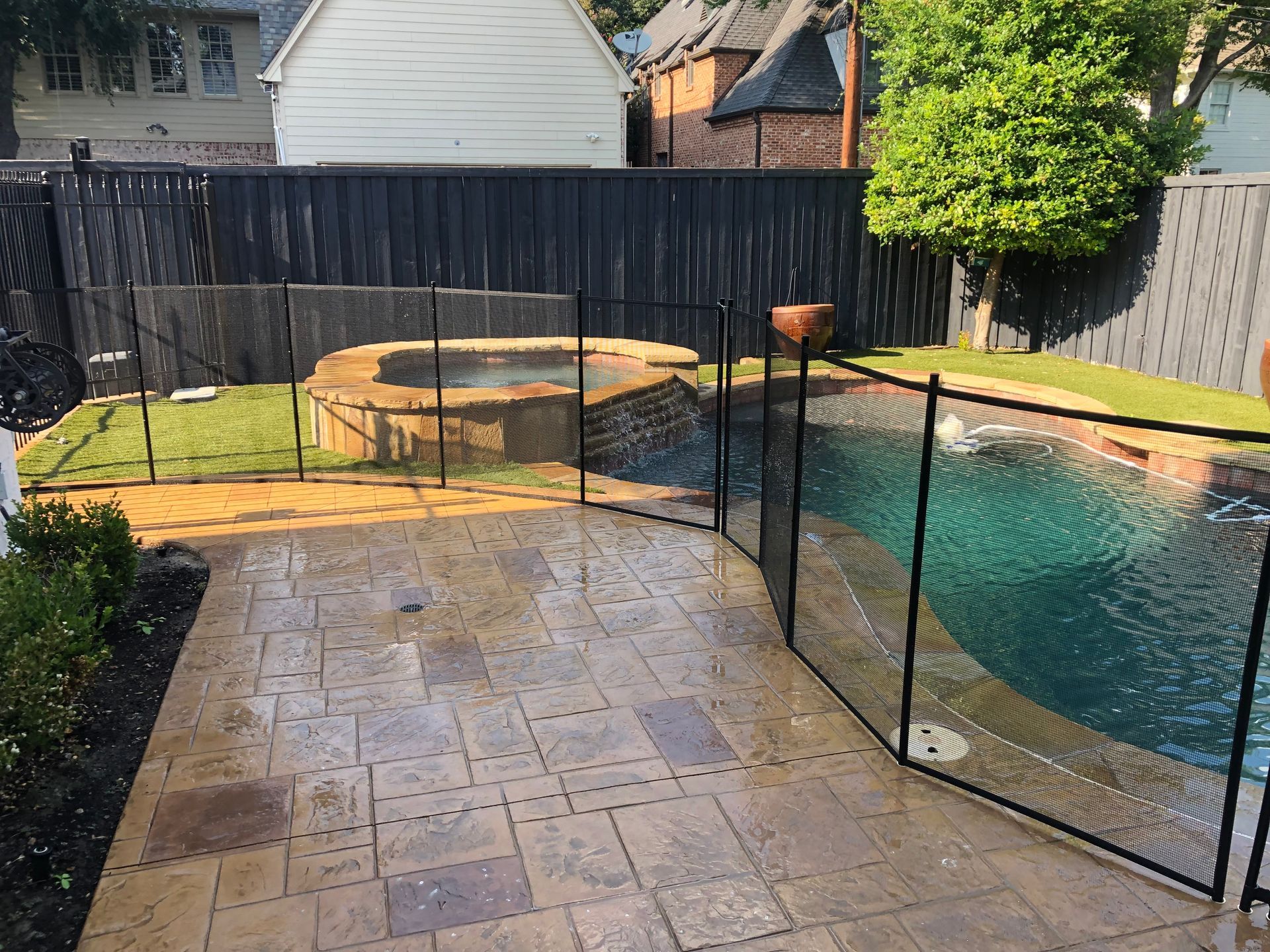 Backyard patio with stone pavers, black safety fence, and a swimming pool beside a hot tub
