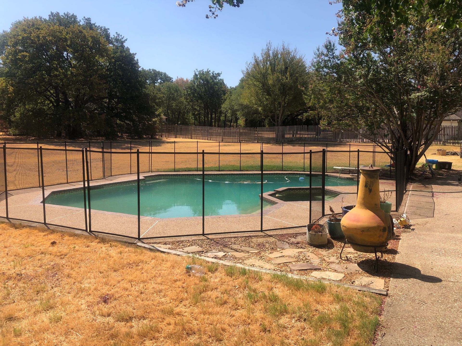 Fenced backyard pool with turquoise water, surrounded by dry grass and trees on a sunny day