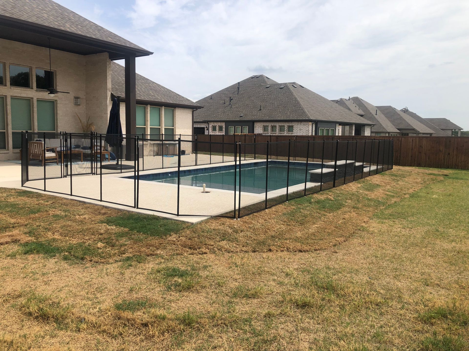 Backyard patio with a fenced swimming pool beside a row of suburban houses and dry grass