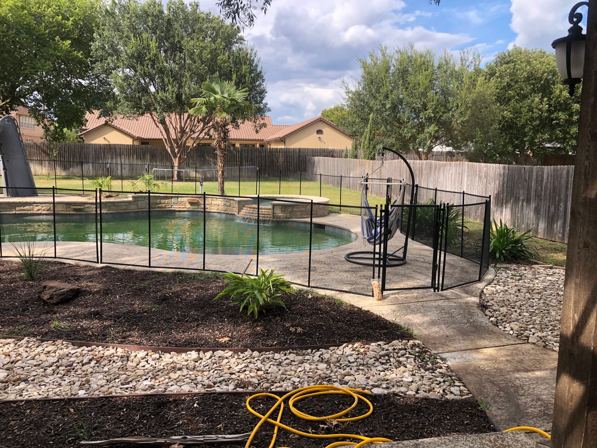 Backyard pool enclosed by black safety fence, with patio, trees, and a garden hose in the foreground.