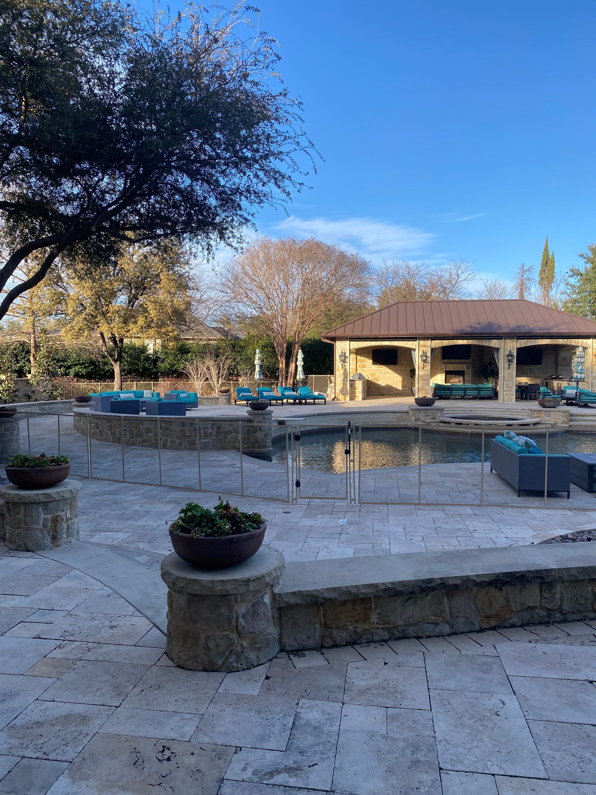 Stone patio overlooking a fenced pool, potted plants, large tree, and a low house in a sunny yard.