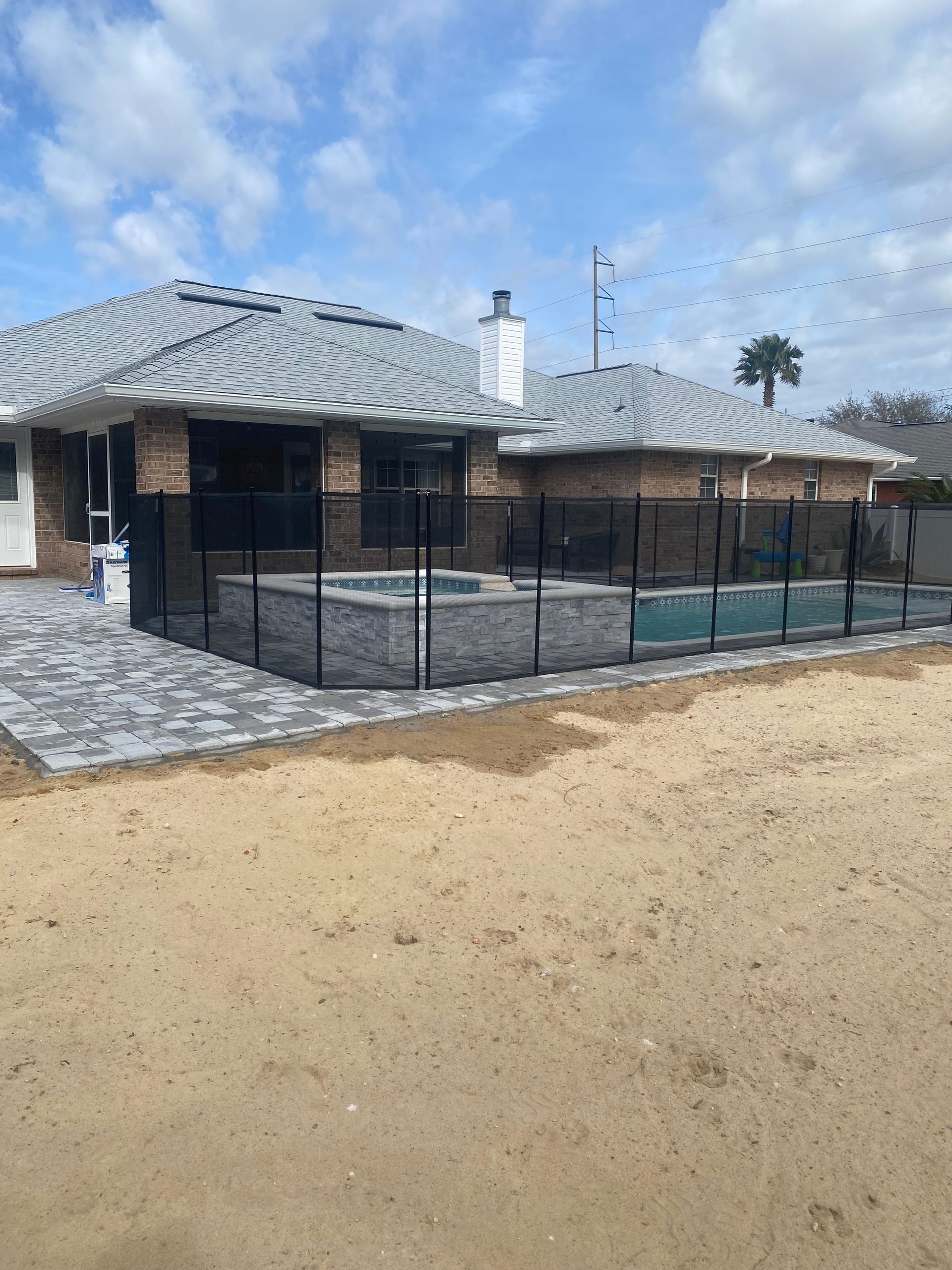 Single-story house with gray tile roof, black pool fence, and sandy yard under a blue sky