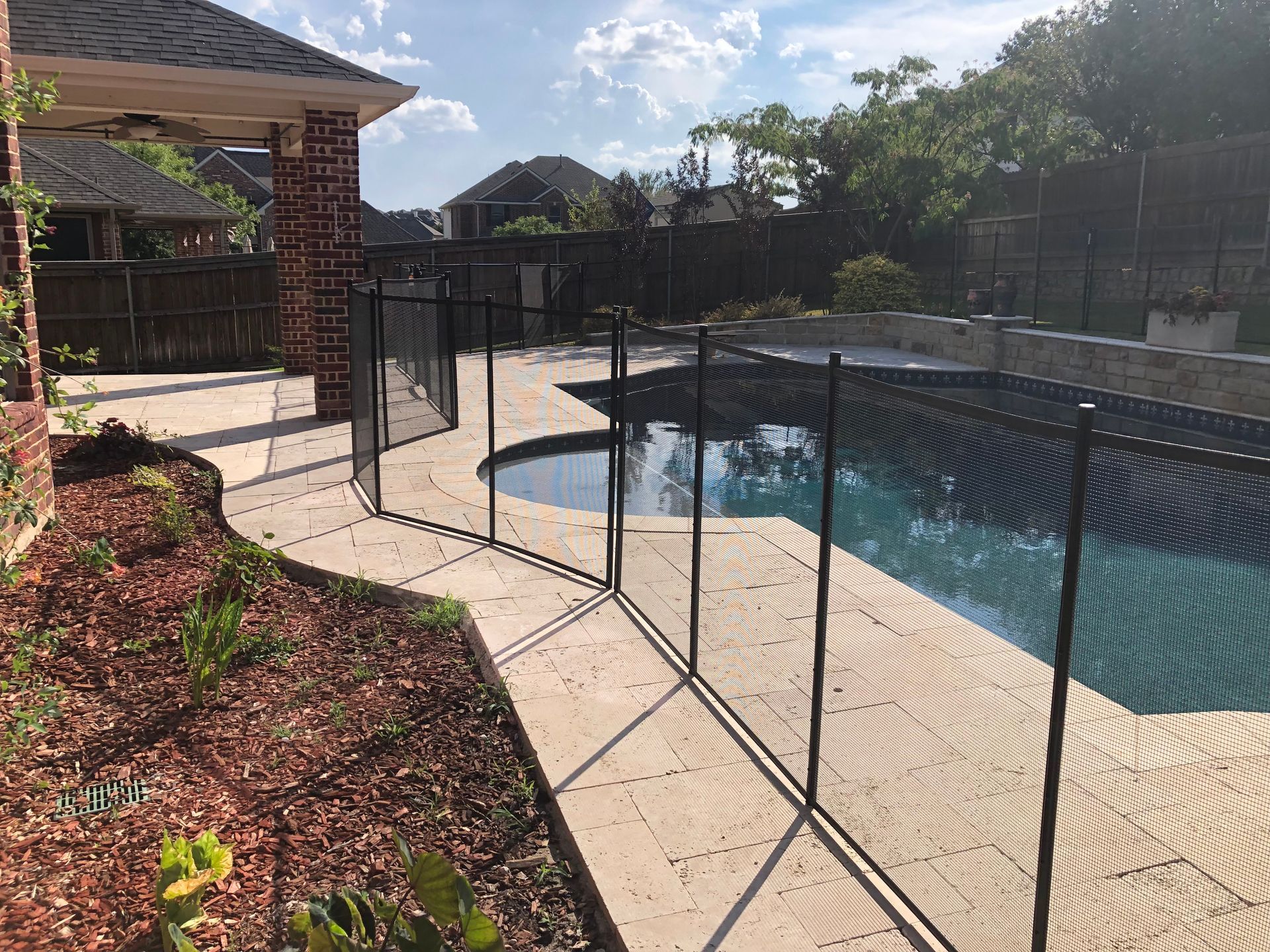 Backyard swimming pool with black safety fence, stone deck, and landscaped garden in sunlight