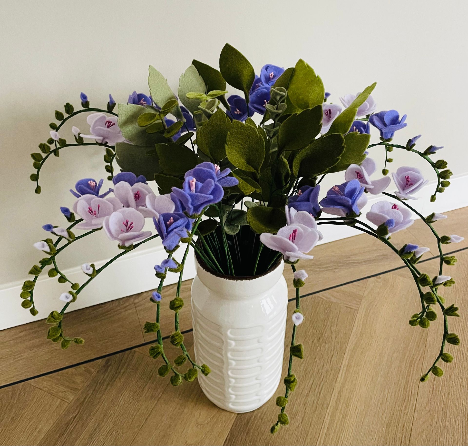 Witte vaas met blauwe en paarse vilten bloemen, groene bladeren op een houten vloer.