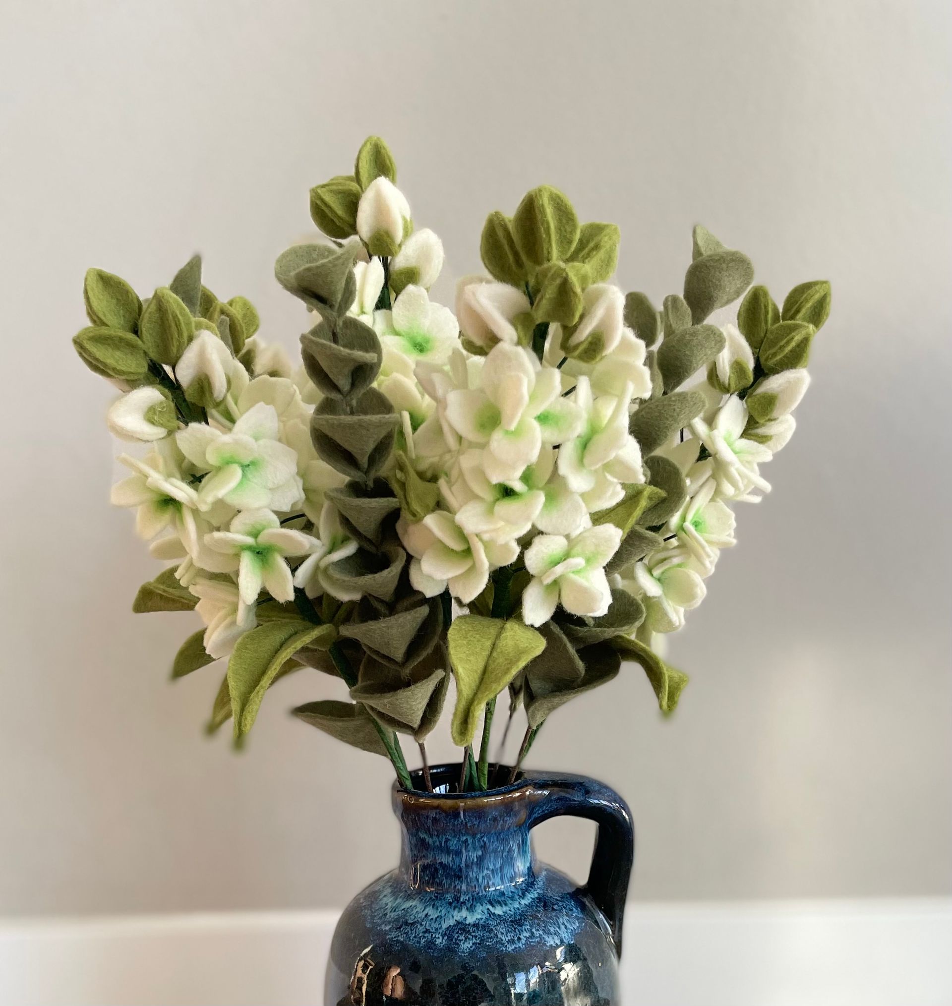 Witte vilten bloemen, stokroos, met groene bladeren in een donkerblauwe keramische vaas.