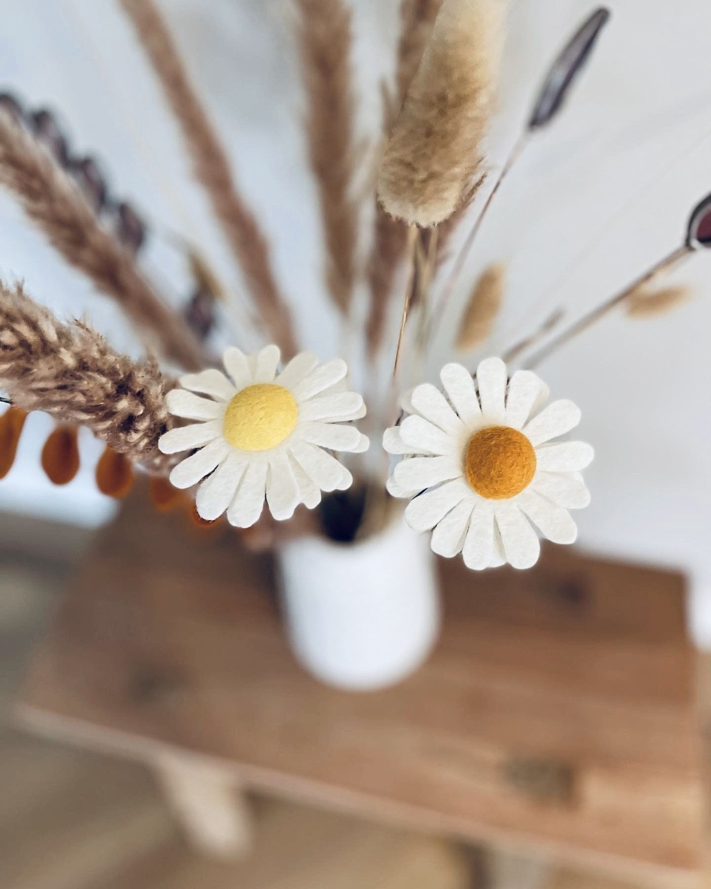 Witte vaas met twee vilten madeliefjes, omringd door gedroogd gras, op een houten krukje.