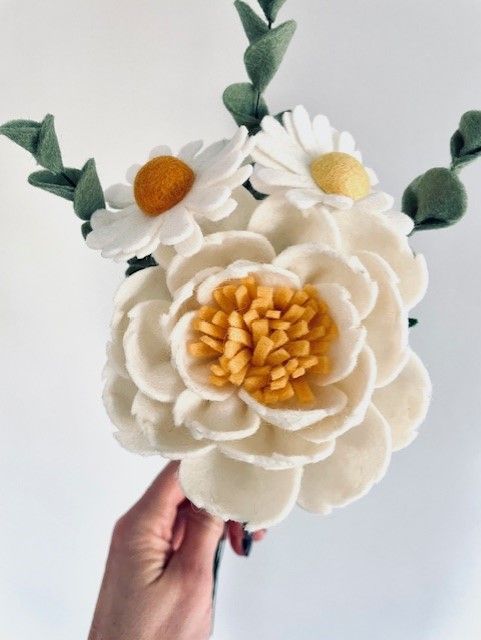 Hand met een boeket vilten bloemen, waaronder witte madeliefjes, een grote witte pioenroos en groene eucalyptus.