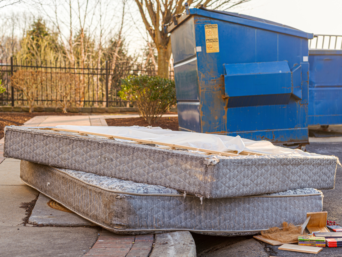 Two discarded, stained mattresses stacked near a blue dumpster in an outdoor setting.